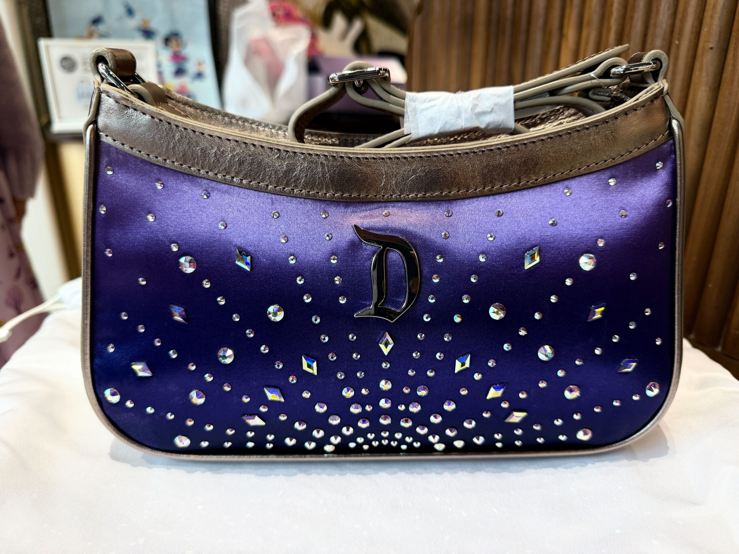 The matching purse is shiny purple with Swarovski crystals on the front. A Disneyland "D" element is at the center of the crystals. It has metallic silver trim and an adjustable crossbody strap.