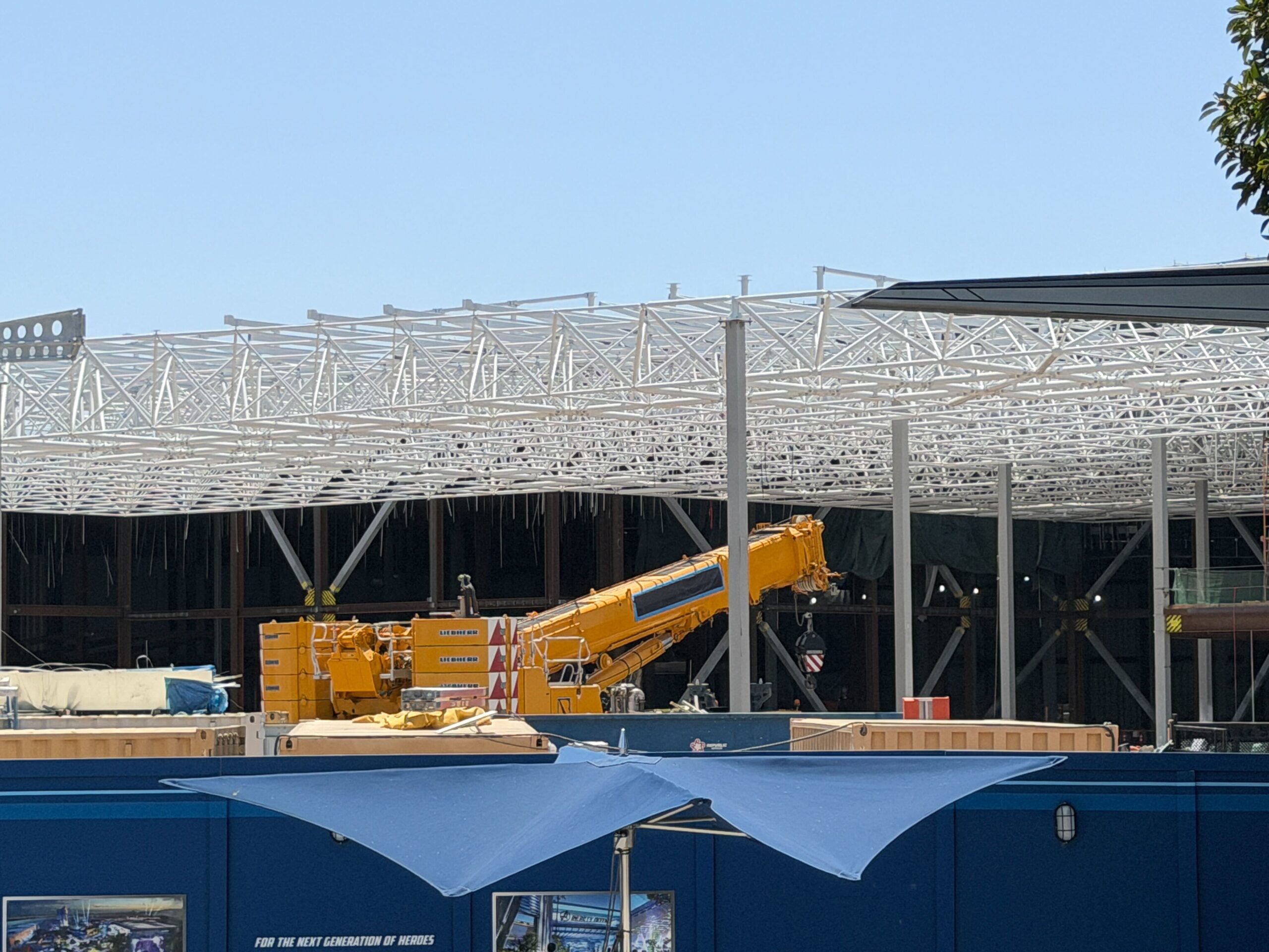 Since our last update, there are fewer boom lifts underneath the largely unchanged, white steel canopy frame, with just one yellow vehicle remaining. The vehicle is surrounded with construction materials.
