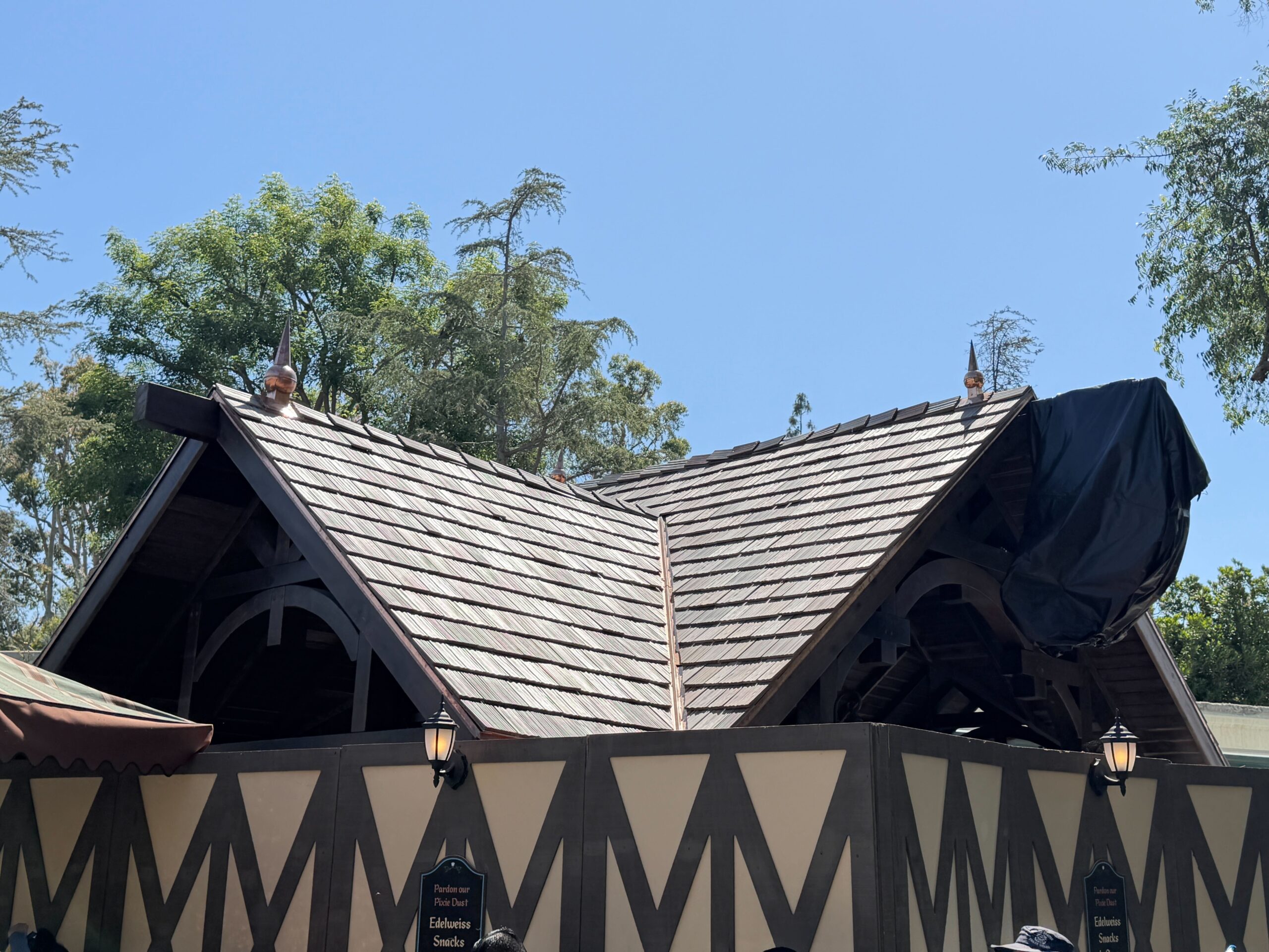 The completed roof, bronze spires, and covered sign can be seen from the other side of the walkway. Construction walls still surround the building and are decorated with lanterns and small signs reading "Pardon Our Pixie Dust: Edelweiss Snacks is Open."