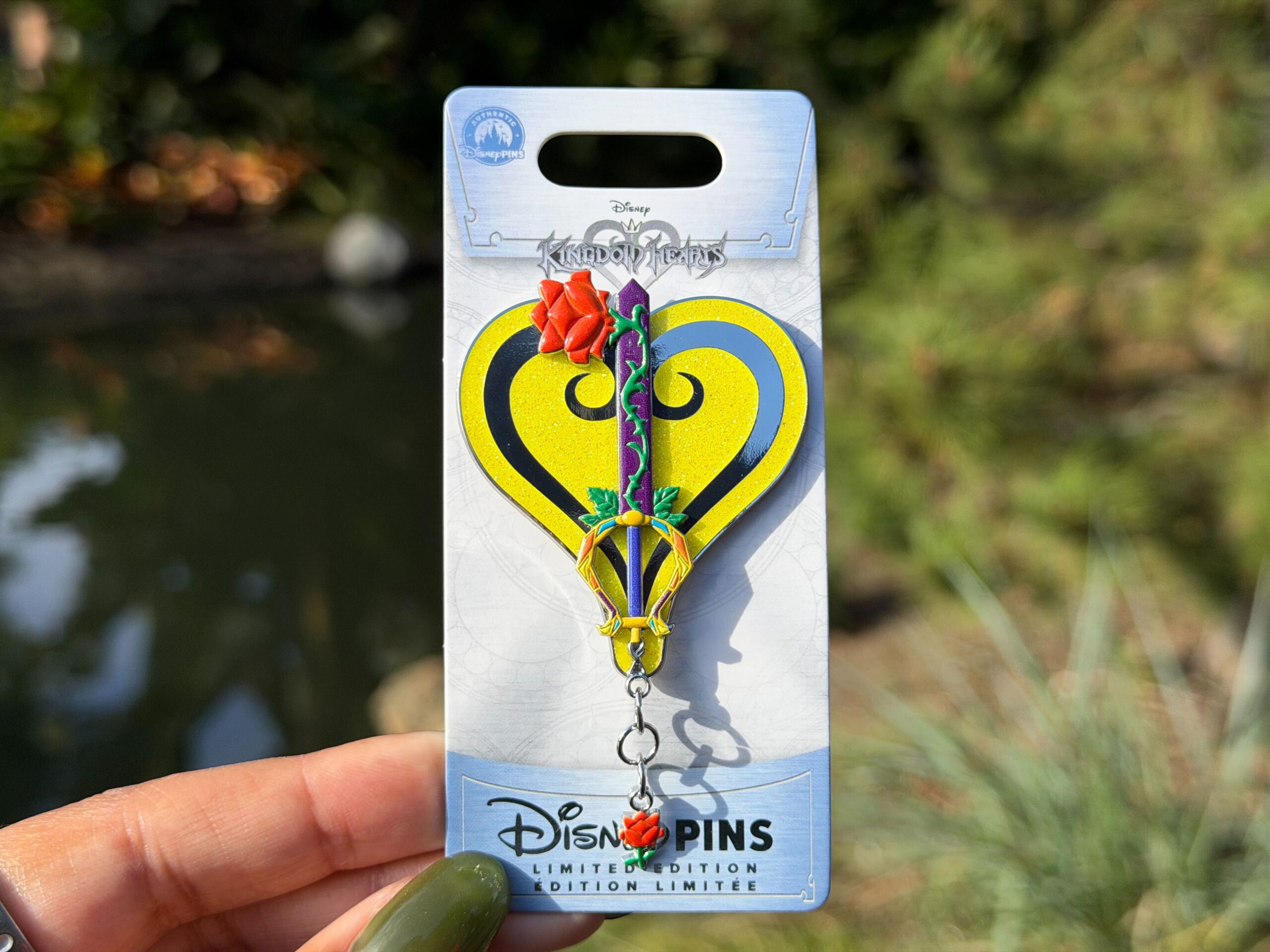 This pin depicts the Beauty and the Beast-inspired Divine Rose Keyblade from Kingdom Hearts. A green thorny stem creeps up the purple blade to a red rose. The handle is yellow and gold. It's set against a sparkly yellow background and a rose charm hangs from the bottom.