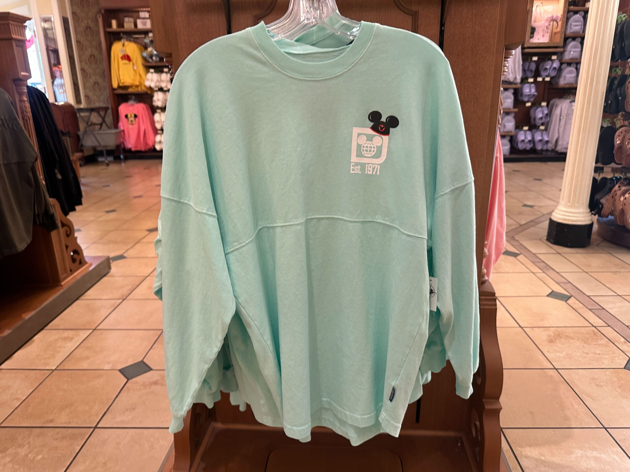 In mint green, this Spirit Jersey shows a white Walt Disney World logo on the lefthand chest, wearing a Mickey ear hat.