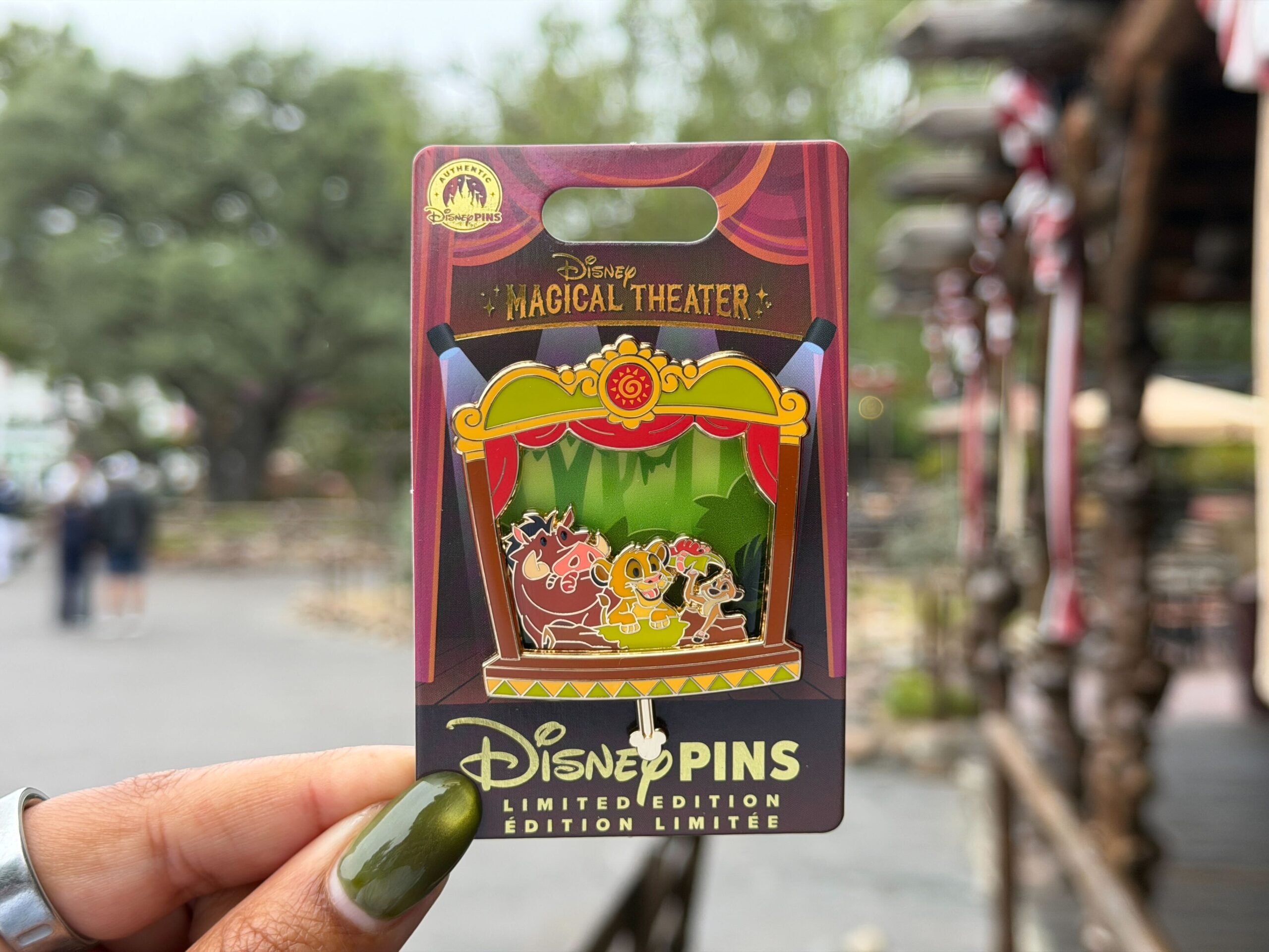This is the fifth monthly "Magical Theater" pin. Simba, Pumbaa, and Timon are pictured within a brown, gold, and green theater proscenium with red curtains. The trio leans on a mossy log with a jungle background.