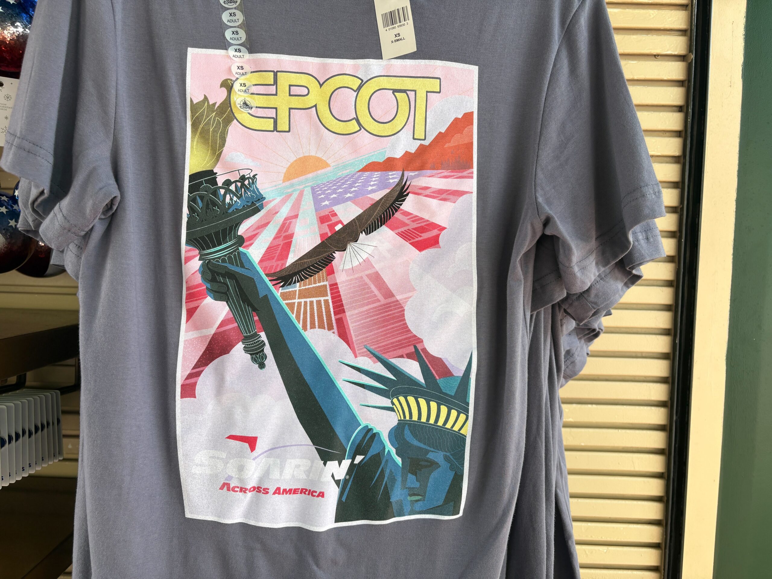 This poster shows the Soarin' Across America logo in the foreground beside a close-up of the Statue of Liberty's head and right arm holding her torch. In the background, we see the American flag superimposed over farm land from a bird's eye view. Across the flag is a bald eagle taking flight towards the sun on the horizon.