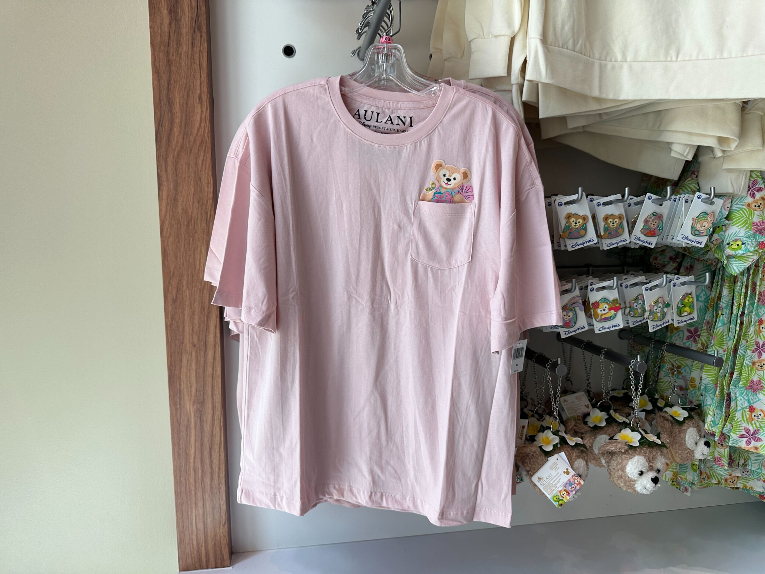 The tee is pale pink with a chest pocket. Duffy is pictured peeking out, wearing a blue and pink Hawaiian shirt with flowers behind him.