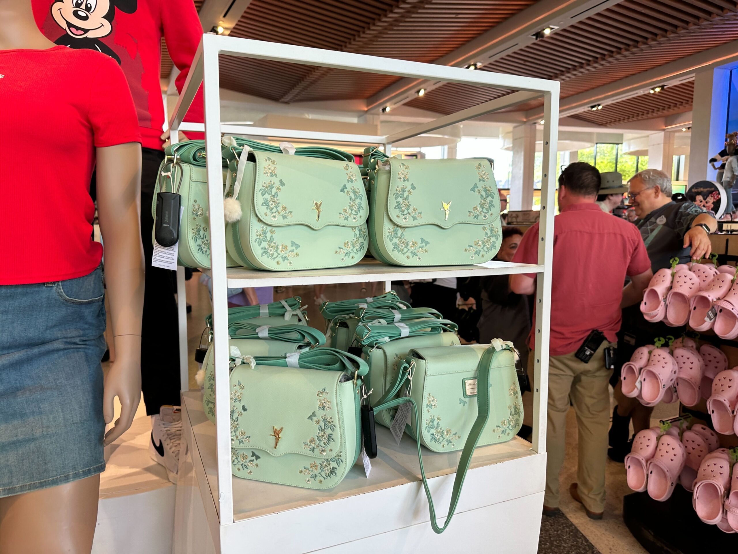 The bag is a light seafoam green color with a scalloped front flap. On each side are embroidered flowers in green, white, and gold colors. In the center is a gold-tone Tinker Bell charm.