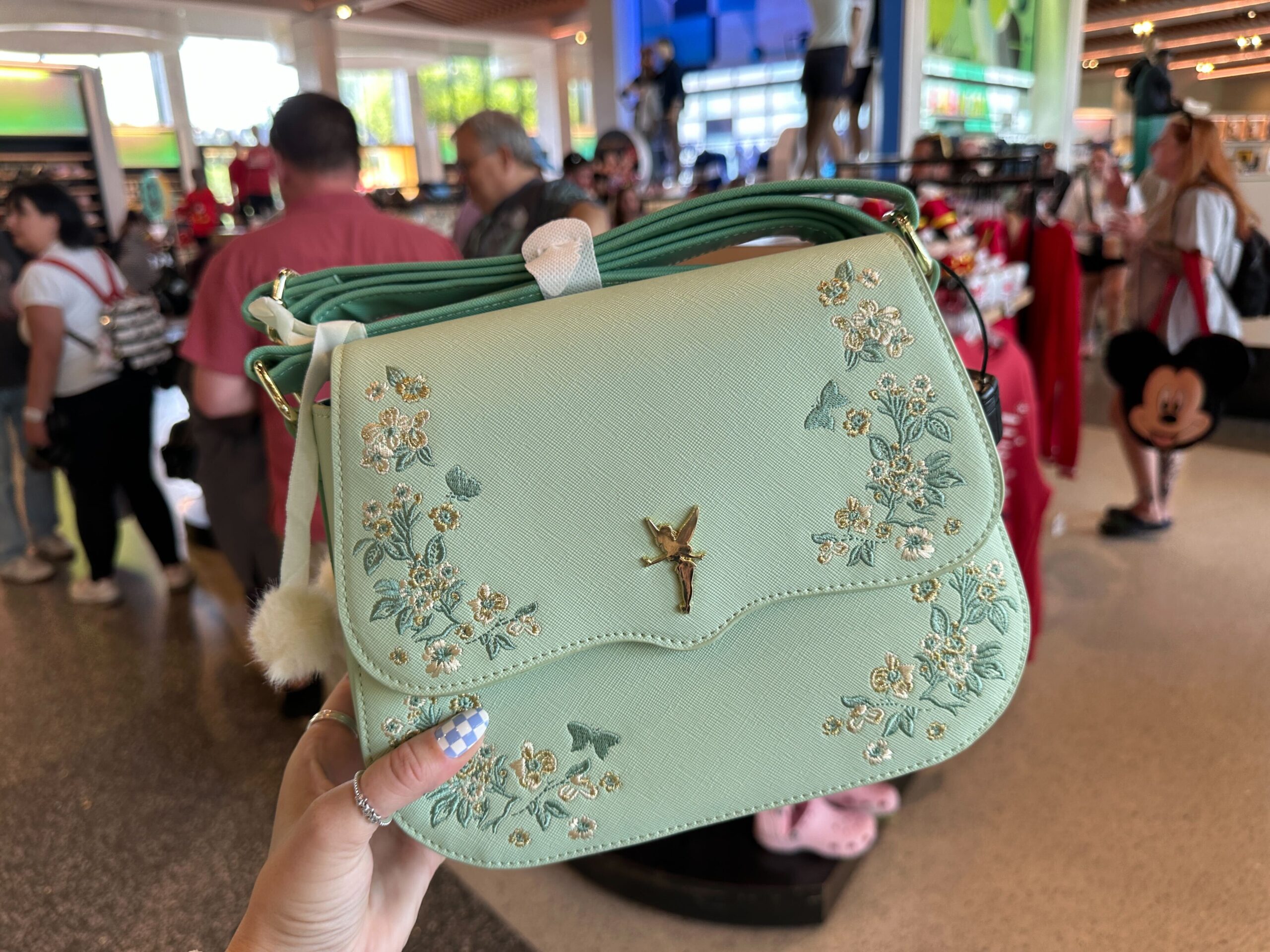 The bag is a light seafoam green color with a scalloped front flap. On each side are embroidered flowers in green, white, and gold colors. In the center is a gold-tone Tinker Bell charm.