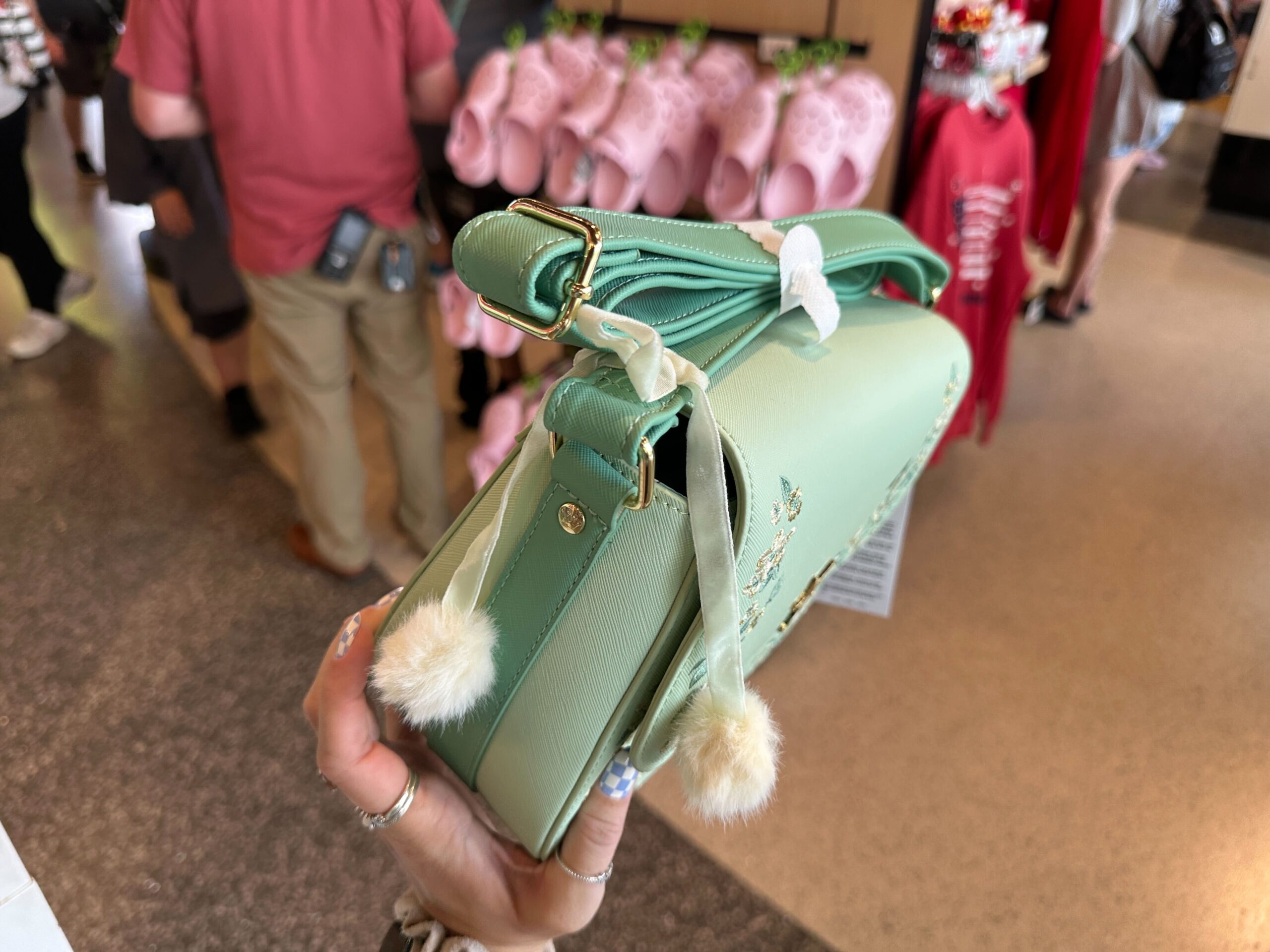 The bag is a light seafoam green color with a scalloped front flap. On each side are embroidered flowers in green, white, and gold colors. In the center is a gold-tone Tinker Bell charm.