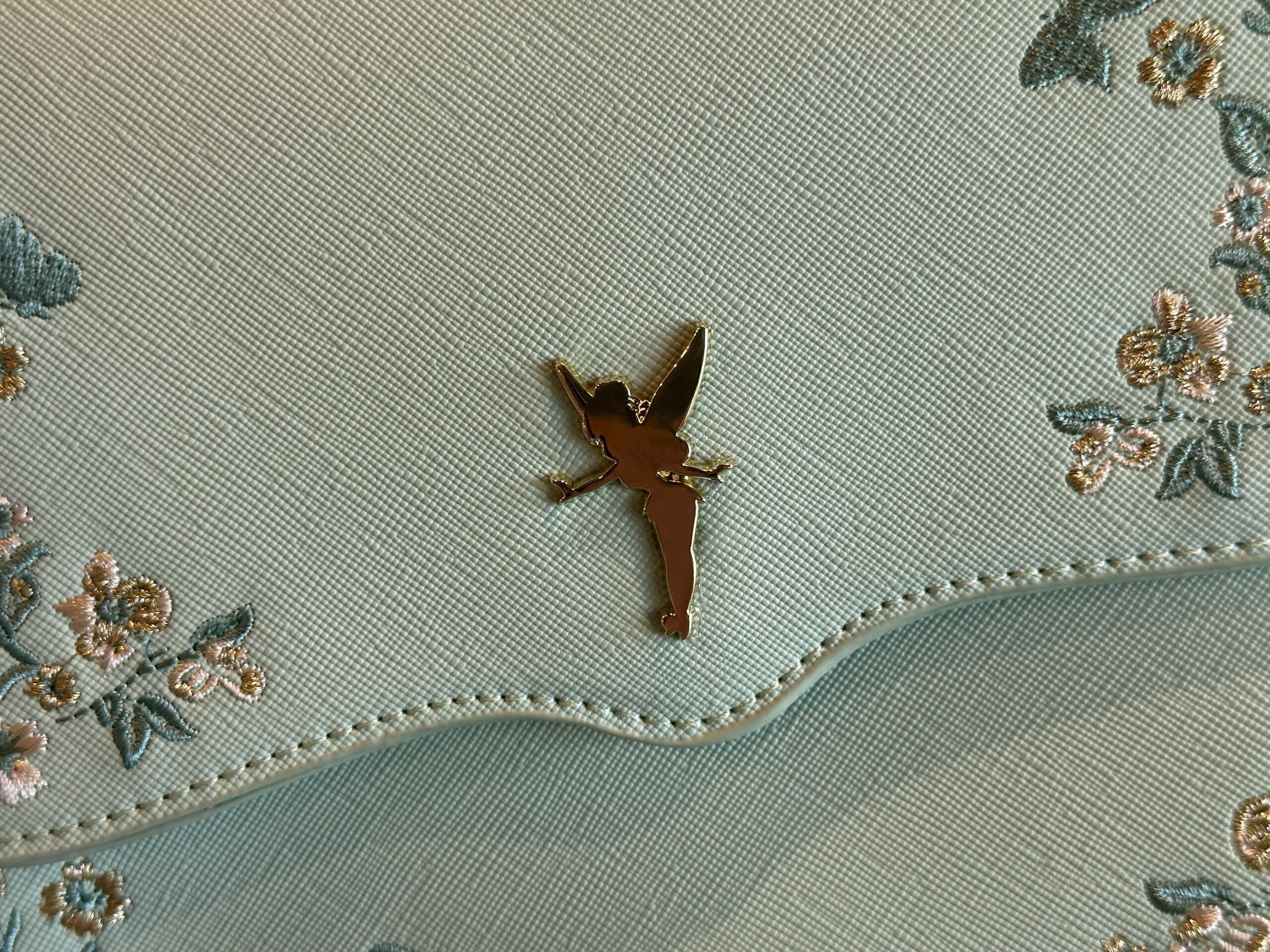 The bag is a light seafoam green color with a scalloped front flap. On each side are embroidered flowers in green, white, and gold colors. In the center is a gold-tone Tinker Bell charm.