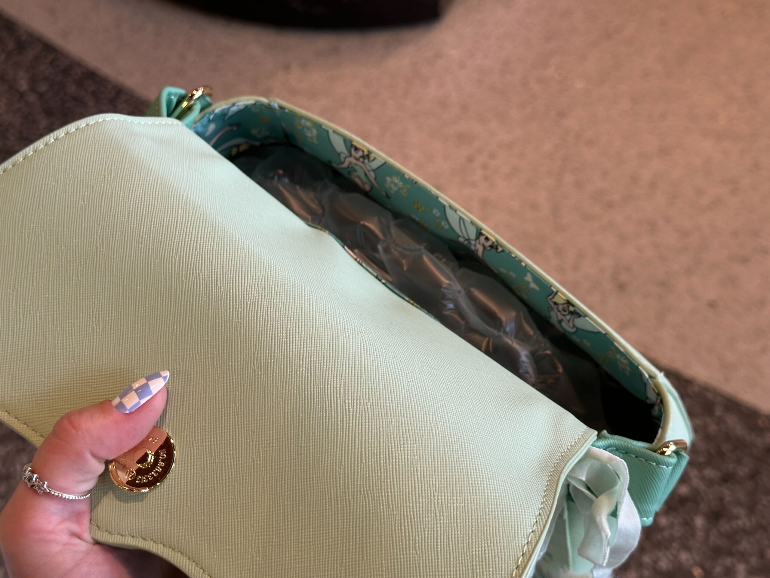 The bag is a light seafoam green color with a scalloped front flap. On each side are embroidered flowers in green, white, and gold colors. In the center is a gold-tone Tinker Bell charm.
