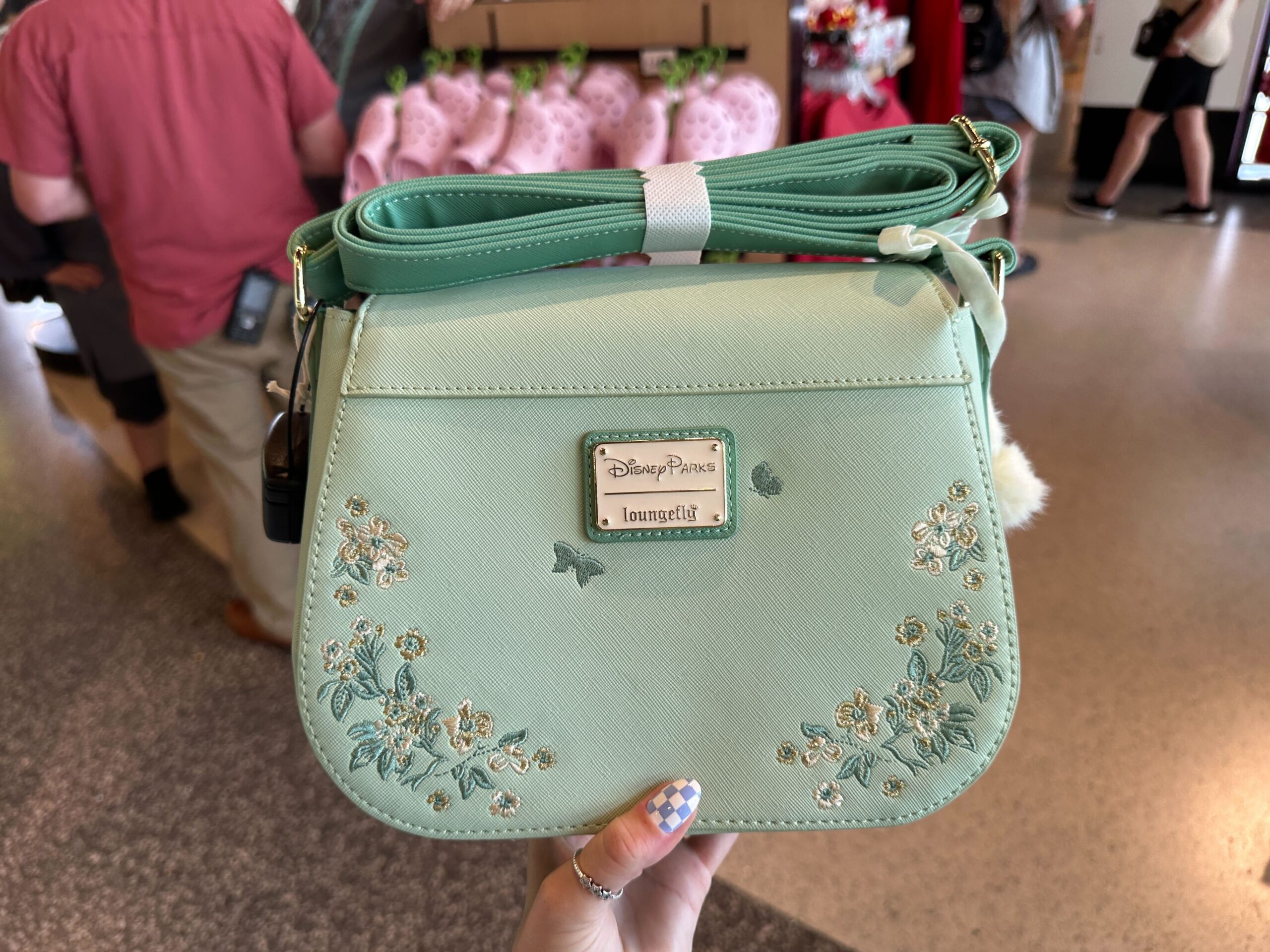 The bag is a light seafoam green color with a scalloped front flap. On each side are embroidered flowers in green, white, and gold colors. In the center is a gold-tone Tinker Bell charm.