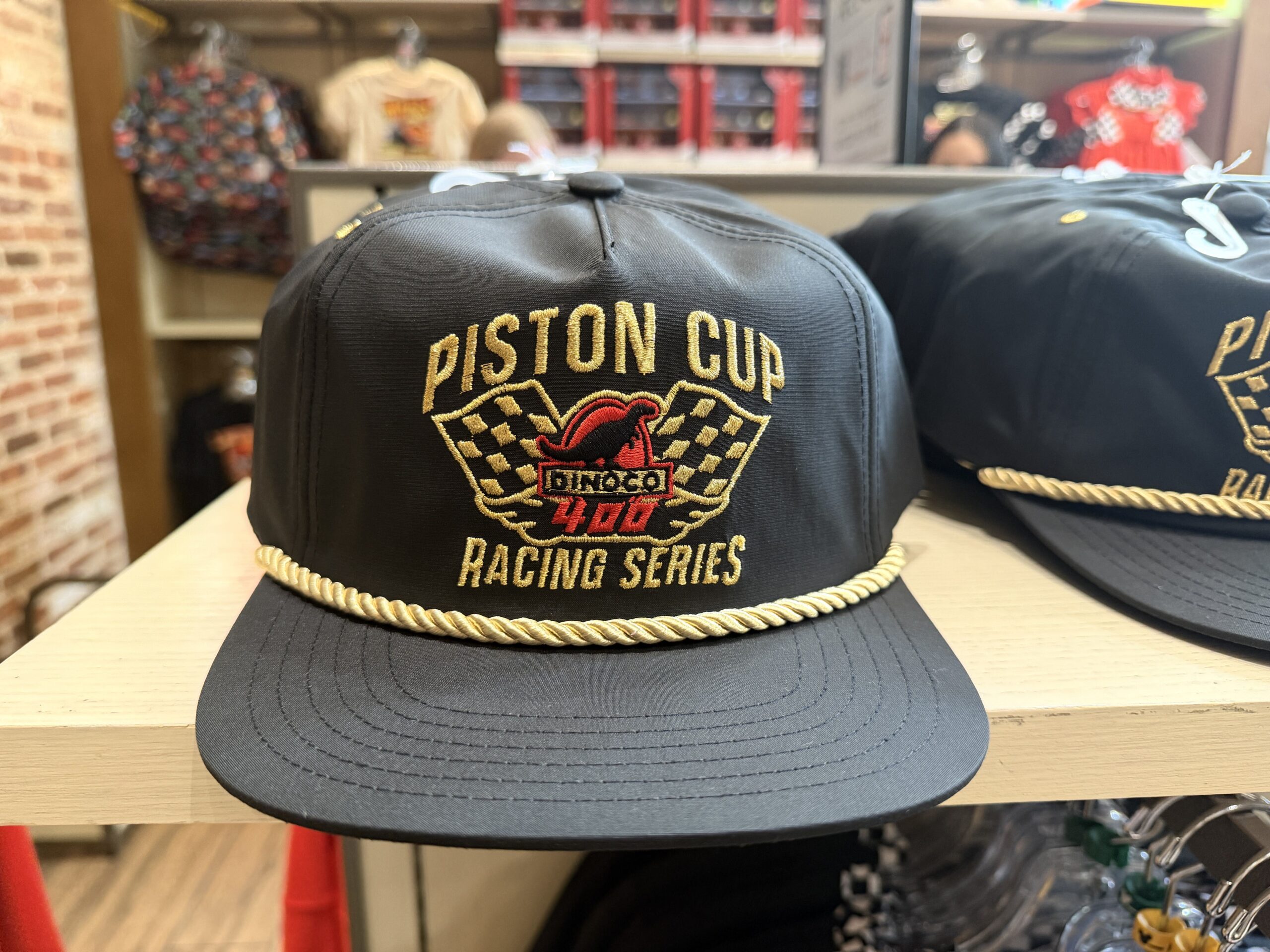 In black, this flat brim cap has a small white rope dividing the space between the brim and the top of the hat. On the front of the hat is white embroidery that reads "Piston Cup" and "Racing Series." In between the set of words is the Dinoco 400 logo in red, white, and black.