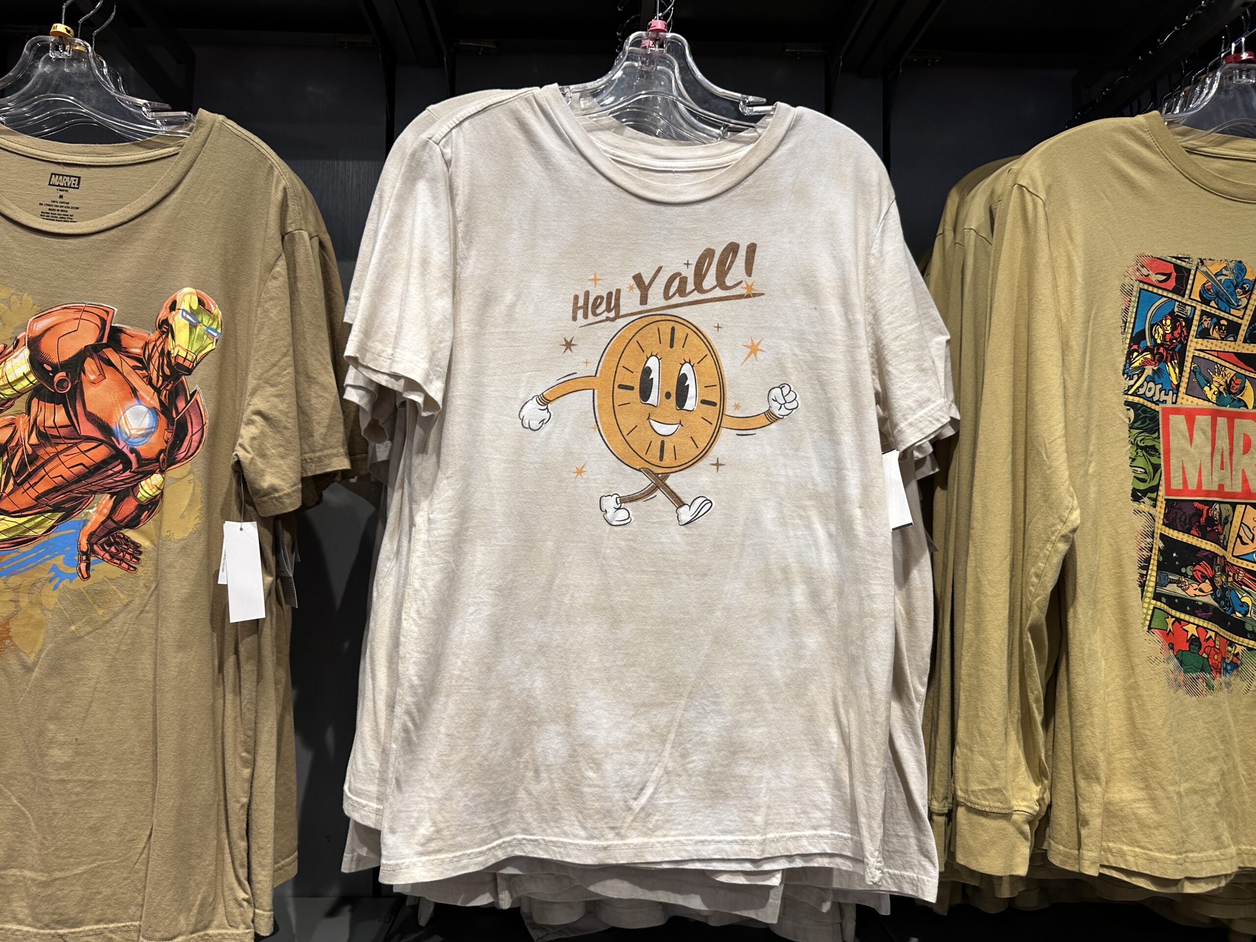 In a tie-dye tan and white, Miss Minutes is shown prominently on the center front of this t-shirt. It says "Hey Y'all!" above her head. She is shown in a retro, dulled color scheme with orange and brown stars around her.