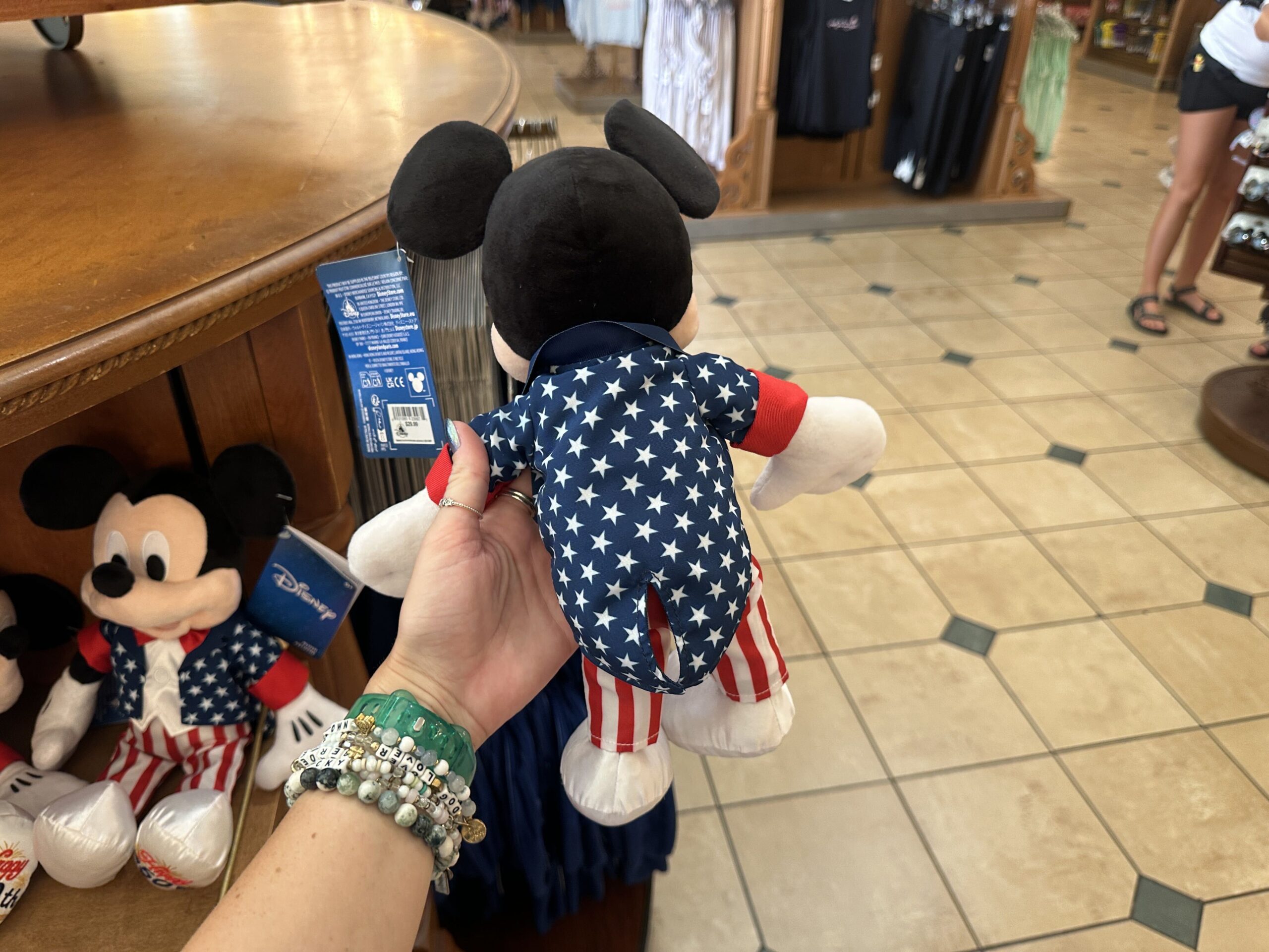 The back of Mickey’s jacket continues the star-spangled pattern across the entire garment, while the striped pants wrap fully around the legs. The plush’s classic smiling expression and outstretched arms give him a welcoming pose that feels ready for Fourth of July festivities.