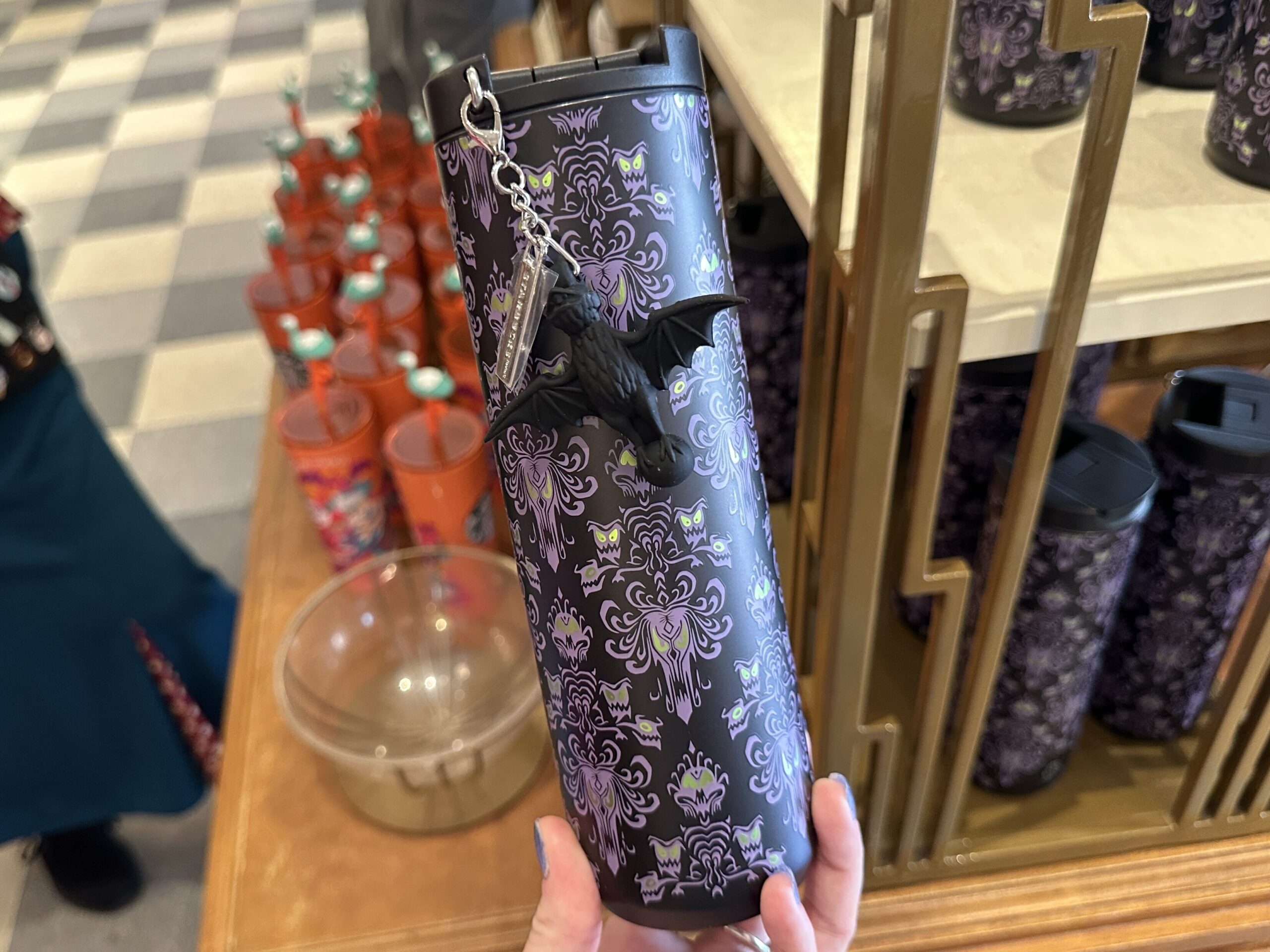 The travel tumbler has a dark, nearly black base color covered in an all-over Haunted Mansion wallpaper-inspired pattern. The design features purple ornamental faces, bats, and eerie green accents that resemble glowing eyes.