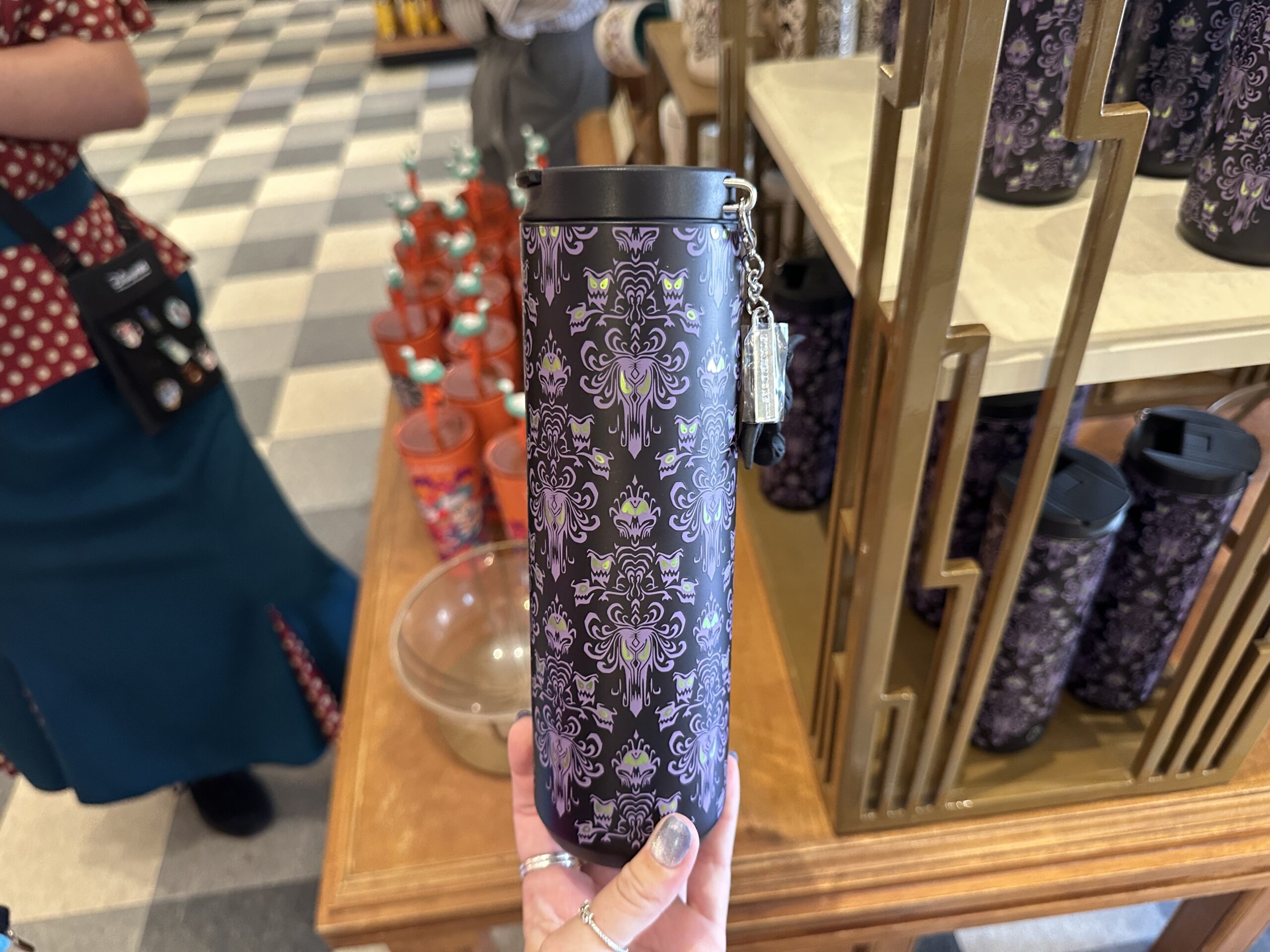 The travel tumbler has a dark, nearly black base color covered in an all-over Haunted Mansion wallpaper-inspired pattern. The design features purple ornamental faces, bats, and eerie green accents that resemble glowing eyes.
