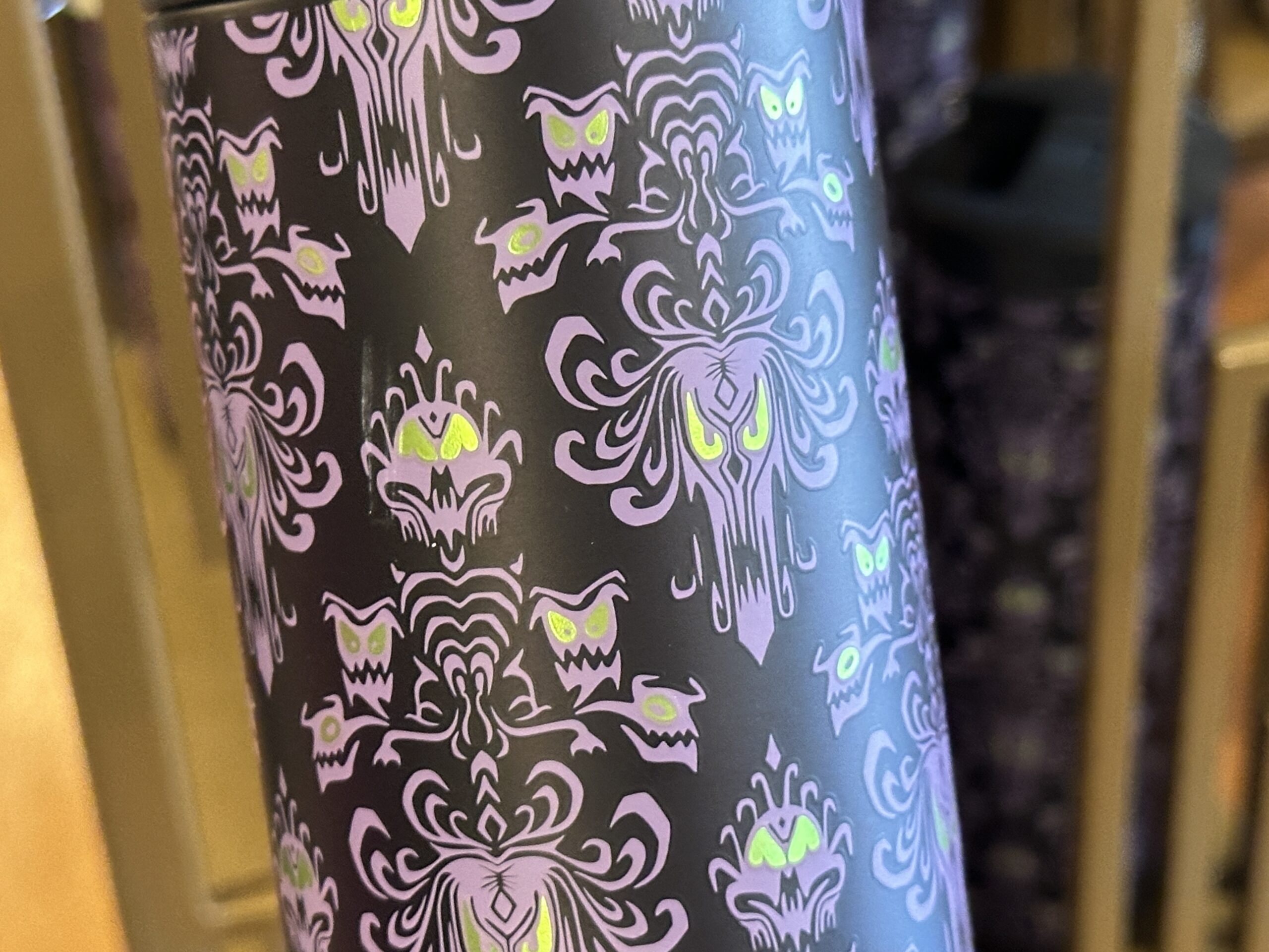 The travel tumbler has a dark, nearly black base color covered in an all-over Haunted Mansion wallpaper-inspired pattern. The design features purple ornamental faces, bats, and eerie green accents that resemble glowing eyes.