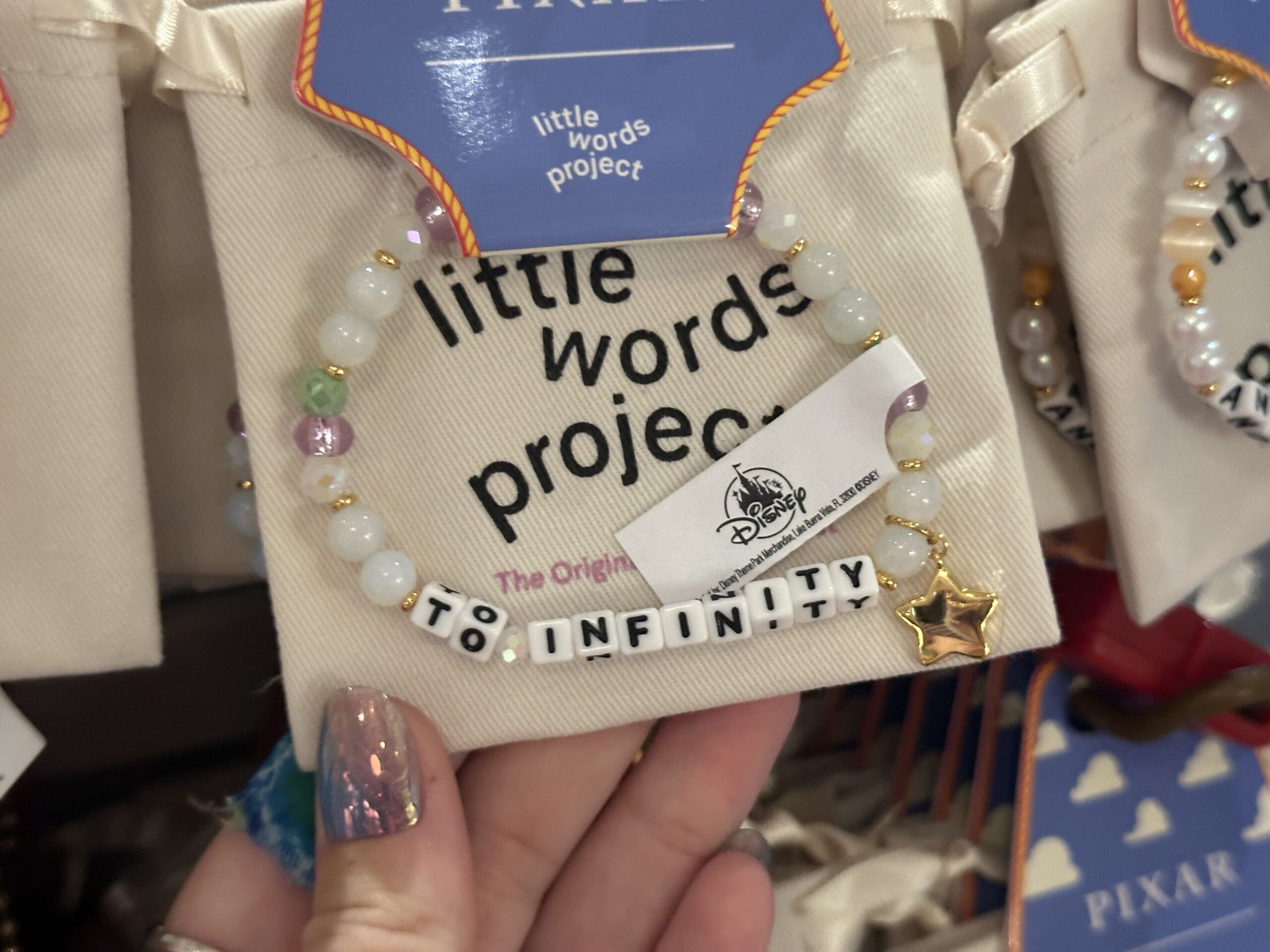 Inspired by Buzz Lightyear's signature catchphrase, this bracelet reads "To Infinity" and features white, purple, and green beads plus a gold-tone dangling star. The full catchphrase is completed by the next bracelet.