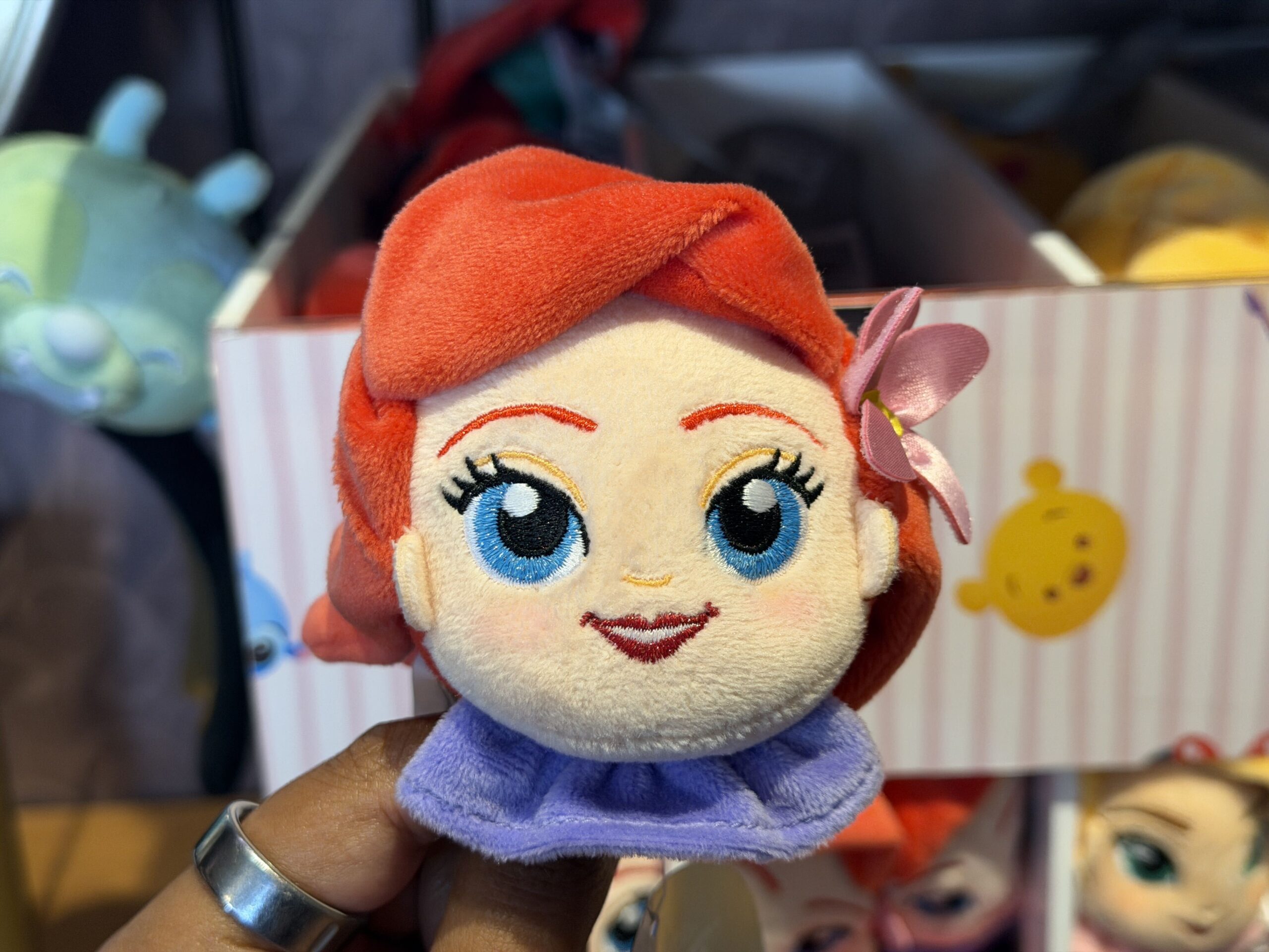 Similar to Rapunzel, Ariel's soft, fabric hair also has a flower. However, this pink flower is a fabric element that sticks out from behind her left ear. Her facial features are embroidered and she has a purple ruffle beneath her face.