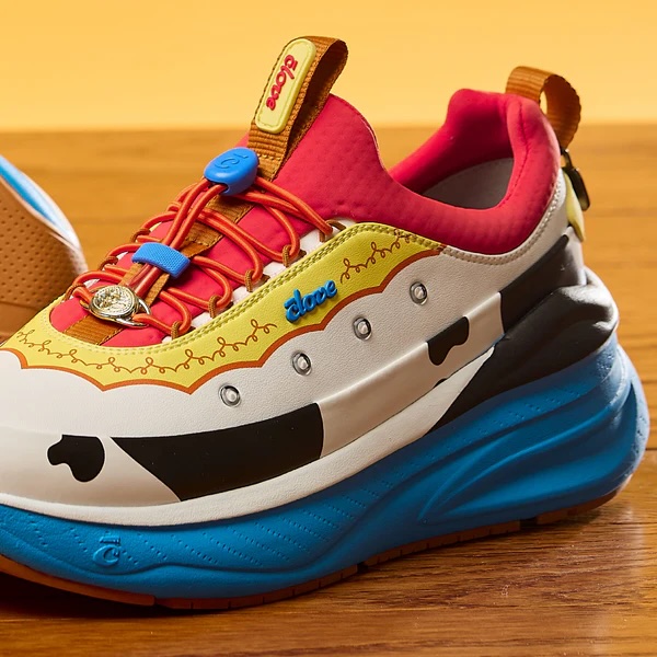 The Jessie shoes are red on top, followed by yellow printed trim based on her collar. Jessie's belt buckle is on the laces, and a bow appliqué is on the back. There is a cow print stripe around the side and blue across the bottom.