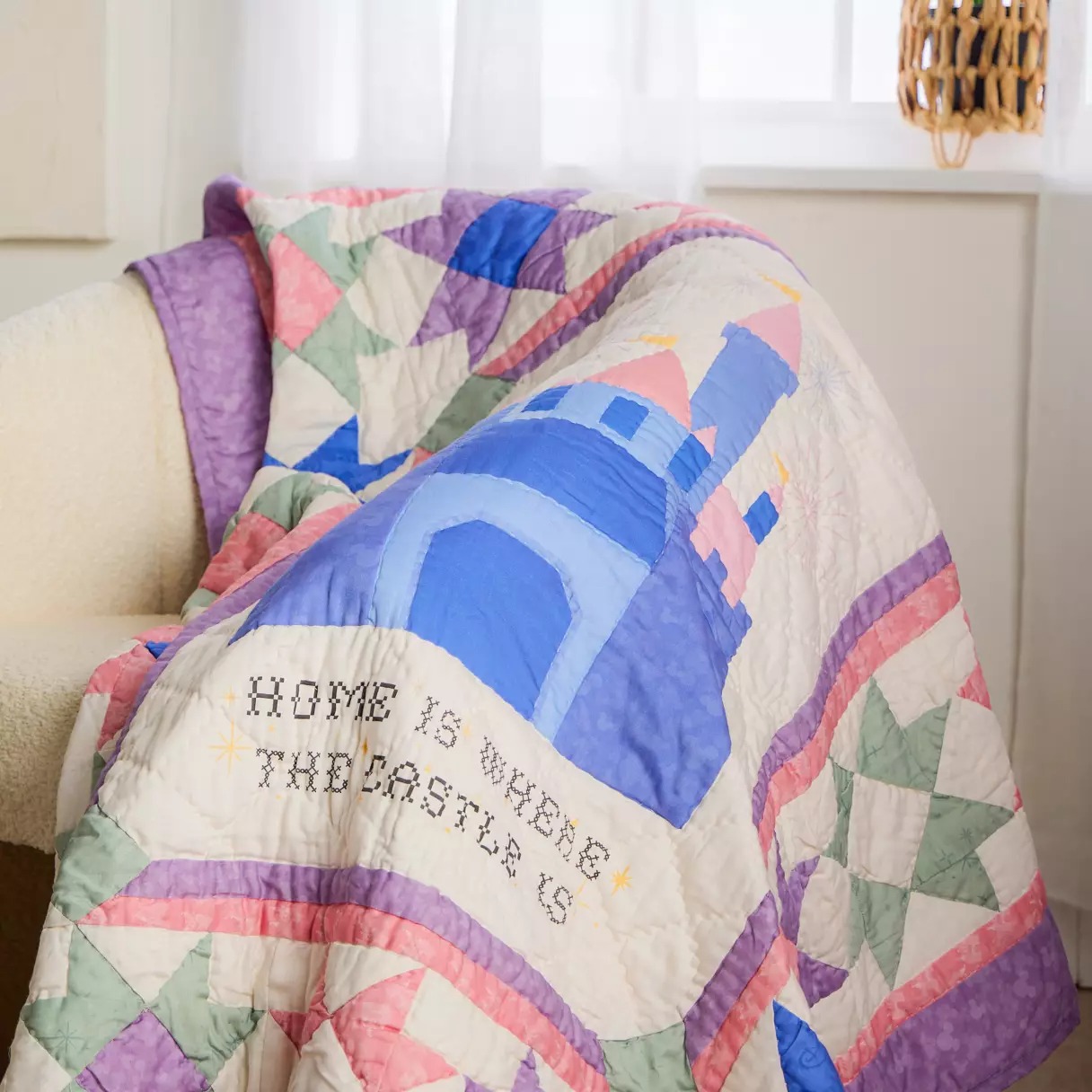 The 100% cotton blanket is 50" x 60." A blue and pink castle is in the center with "Home is where the castle is." It's surrounded by pink, purple, green, and blue quilting stars followed by pink and purple borders, all set against an ivory background.