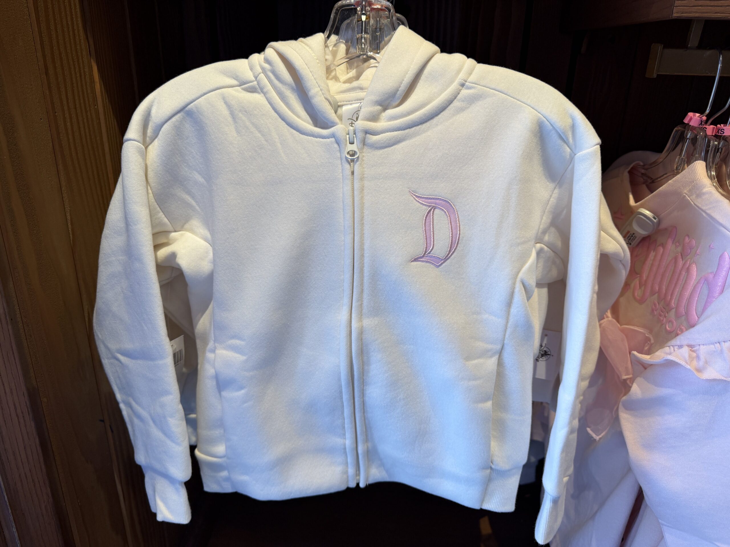 This white zippered youth hoodie has a pink Disneyland "D" on the left front chest. on the back is "Disneyland" in the same shade of pink across the shoulders above a gold outline of Sleeping Beauty Castle.