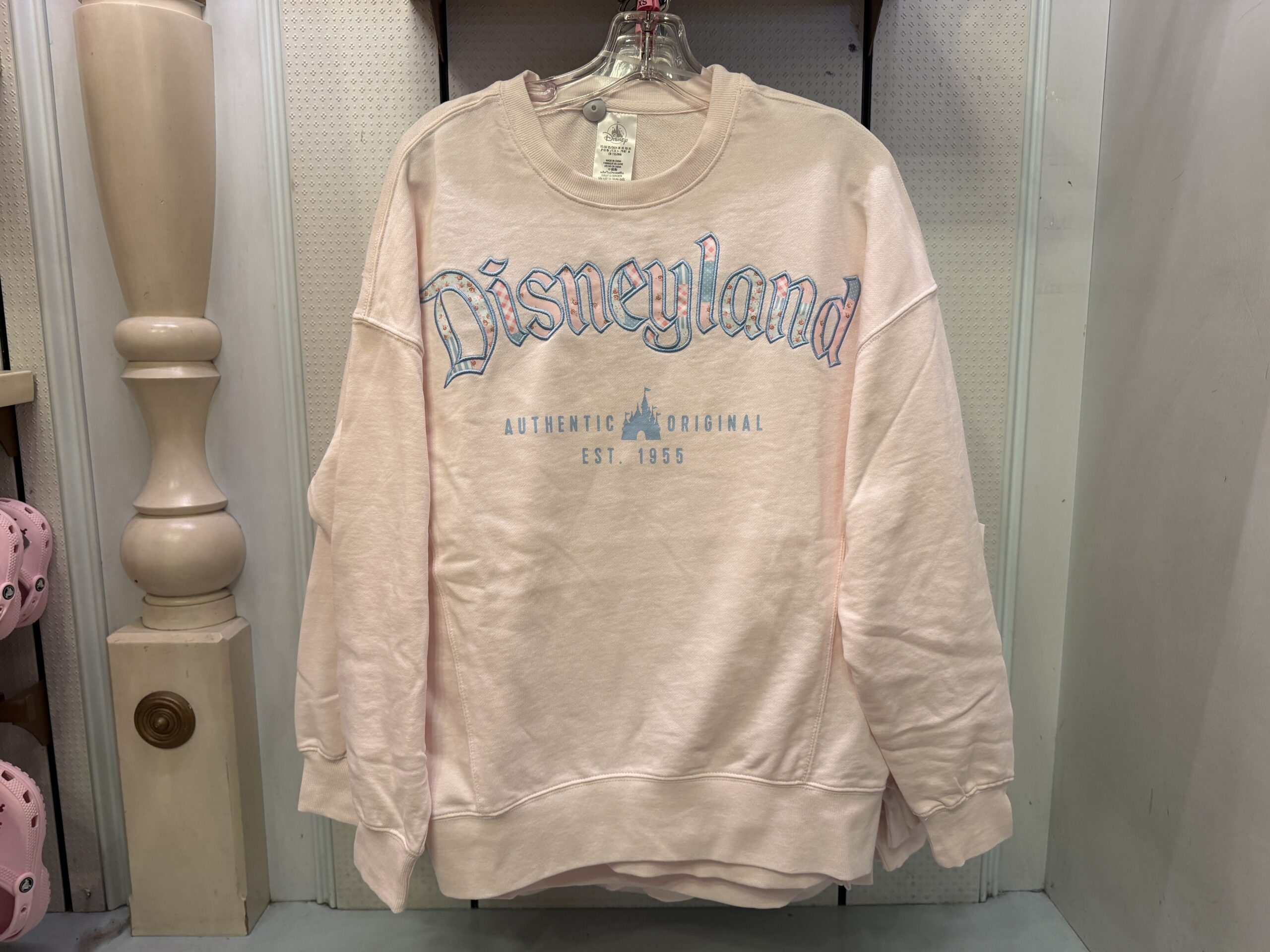 This pullover sweatshirt is pale pink with “Disneyland” across the chest. The wordmark consists of blue and pink contrasting floral, polka-dot, and gingham patterns. The patterned letters are bordered by blue embroidery. Printed below is “Authentic Original Est. 1955” with the castle silhouette, also in blue.