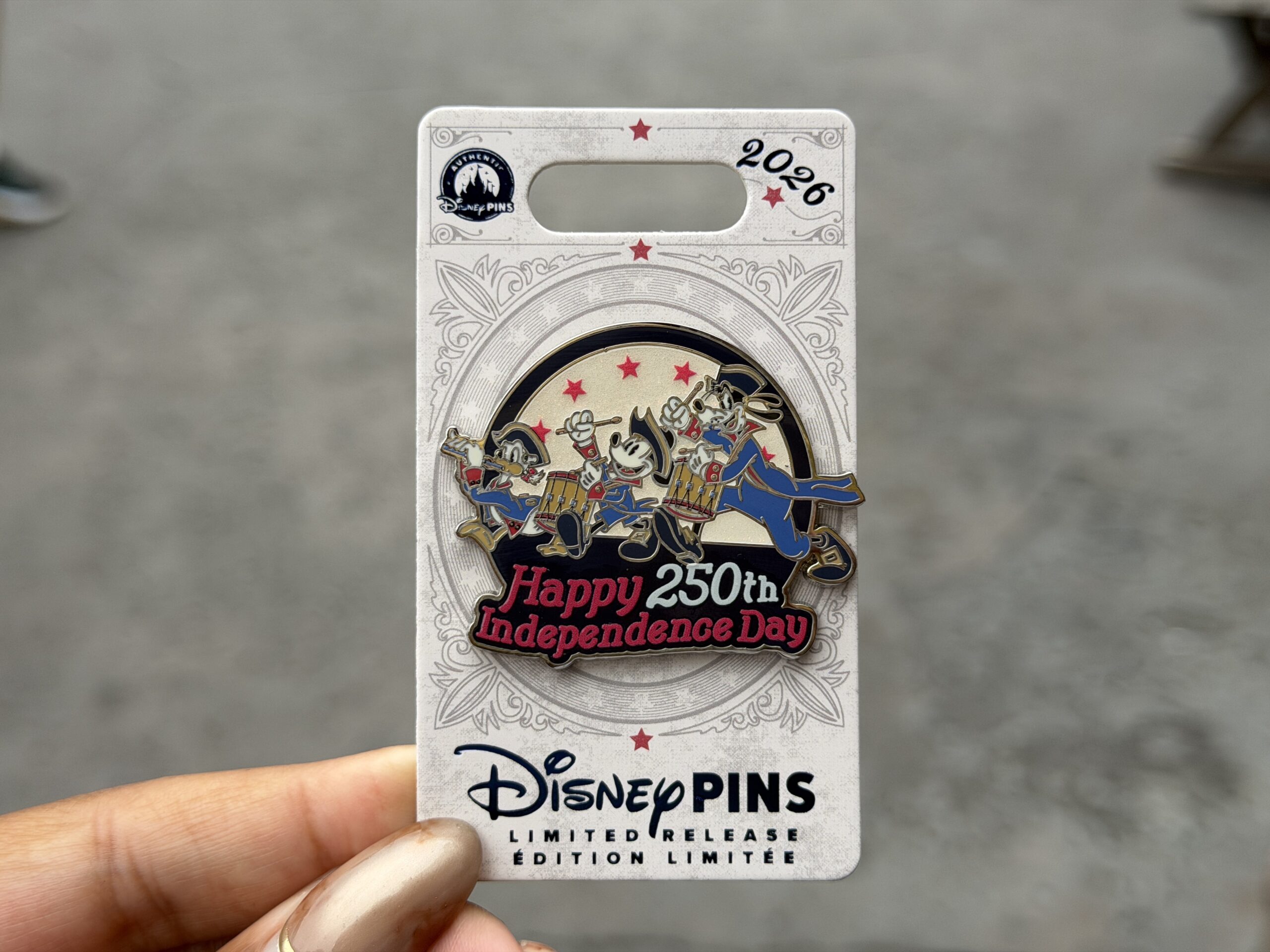 This limited release pin features Donald, Mickey, and Goofy in patriotic uniforms. Donald plays a flute while the other two play drums. "Happy 250th Independence Day" is across the bottom in red and white while stars are in the background.