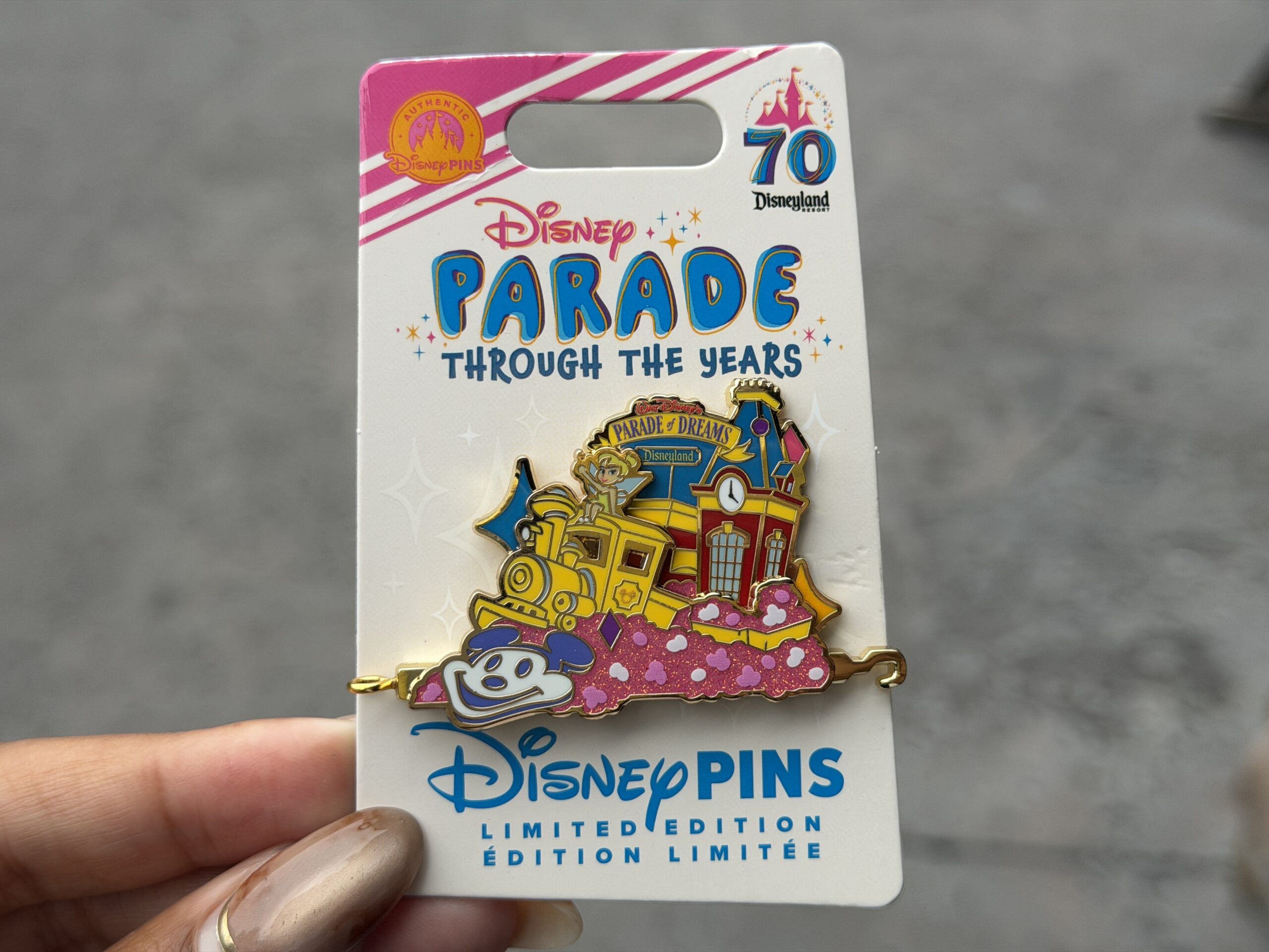 The latest "Parade Through the Years" pin is based on Walt Disney's Parade of Dreams, which celebrated 50 years of Disneyland. The pin depicts the lead float with Tinker Bell atop a train engine and the Main Street train station behind her. Below is a sparkly pink flowerbed featuring Mickey's face.
