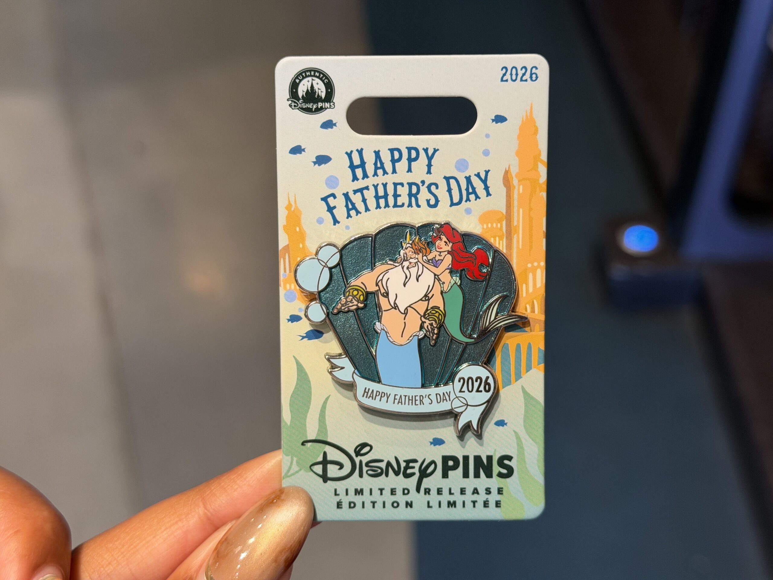 This year's Father's Day pin honors Ariel and King Triton. The princess is placing a flower in her dad's hair. They're set against a sparkly seashell background with bubbles. "Happy Father's Day 2026" is across the bottom.