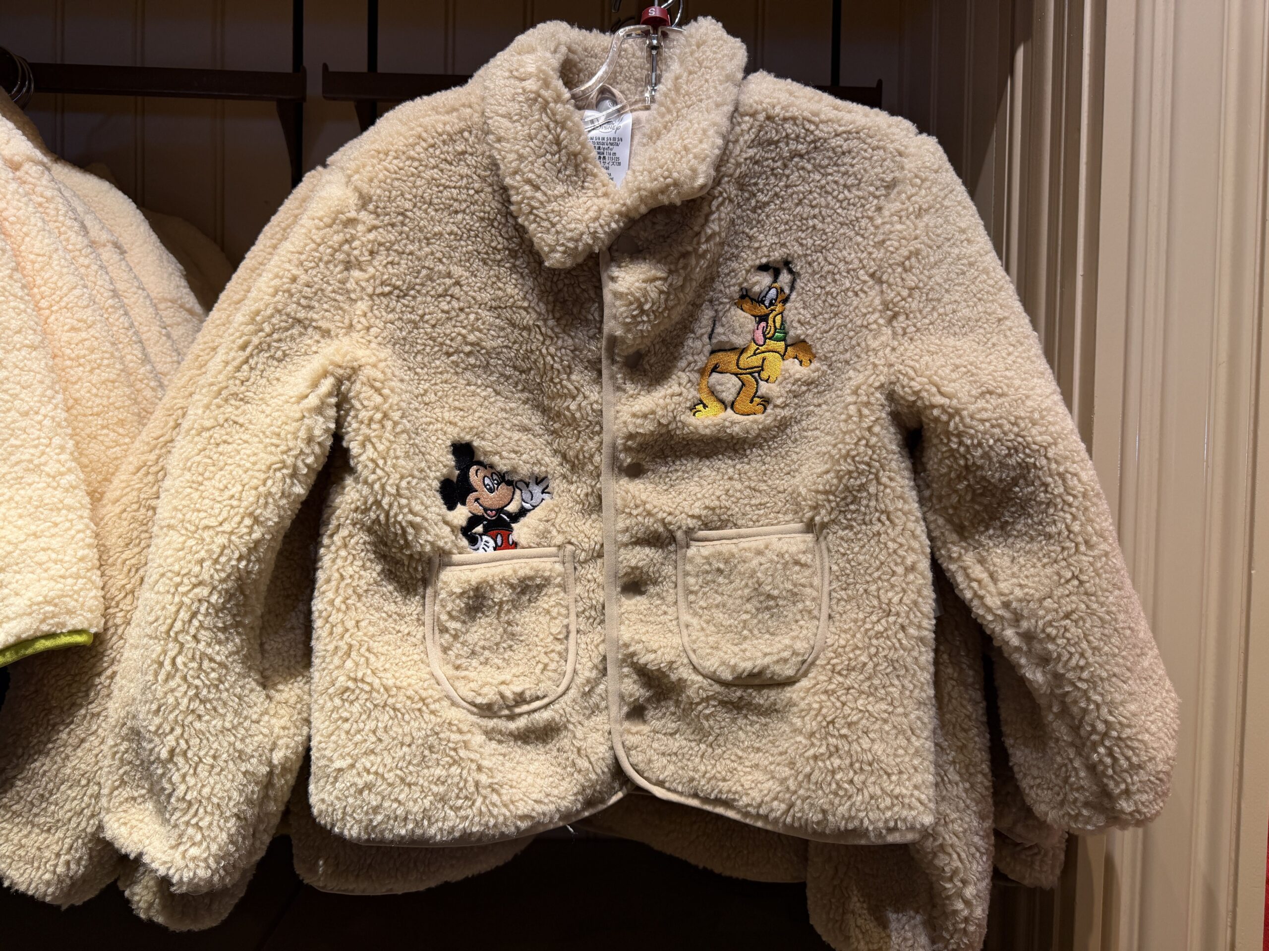 In tan, this sherpa fleece jacket has two front pockets on either side. A patch of Mickey appears to be emerging from the right pocket while Pluto is standing on the left chest.