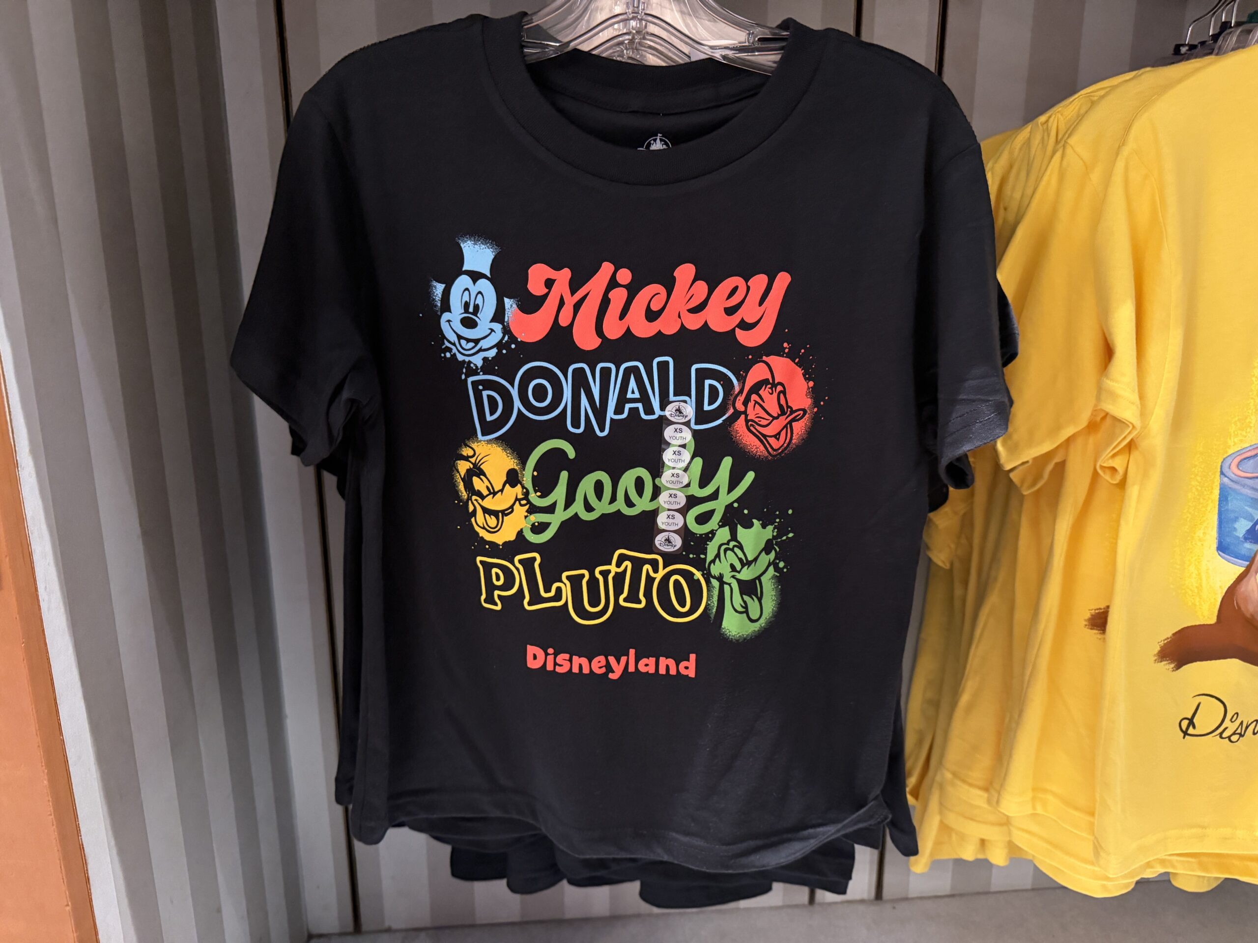 In black, this shirt shows several classic Disney characters' faces in a spray paint circle beside their name. Featured are Mickey, Donald, Goofy, and Pluto. "Disneyland" is in red at the bottom.