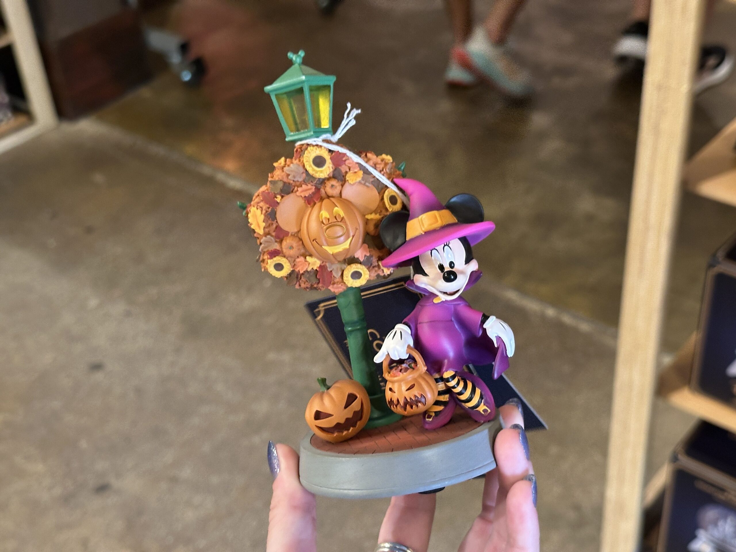 Minnie's witch outfit is purple with orange details. She is wearing orange and black striped leggings with purple heels and holding a Jack-o'-lantern candy bucket.