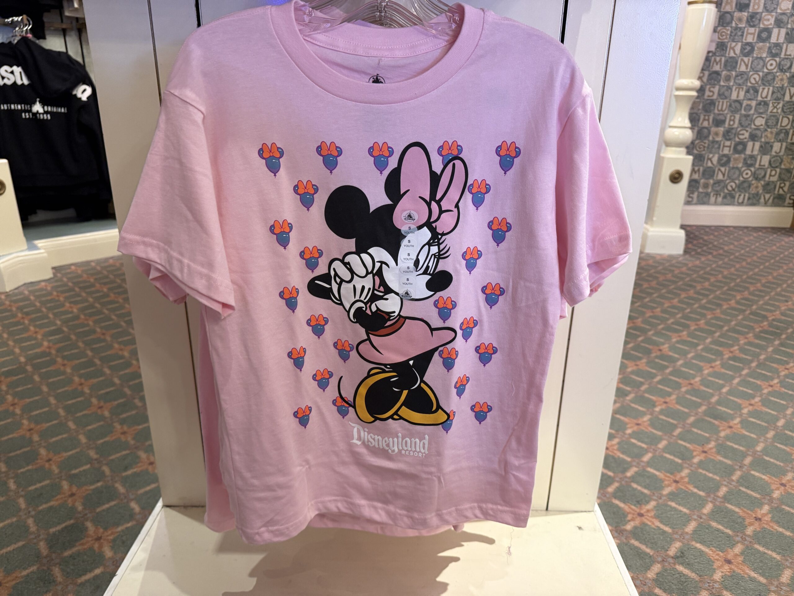 In light pink, this shirt shows Minnie in a classic outfit, posing with her hands by the side of her face. In the background is a pattern of purple Minnie icon-shaped balloons with pink bows.