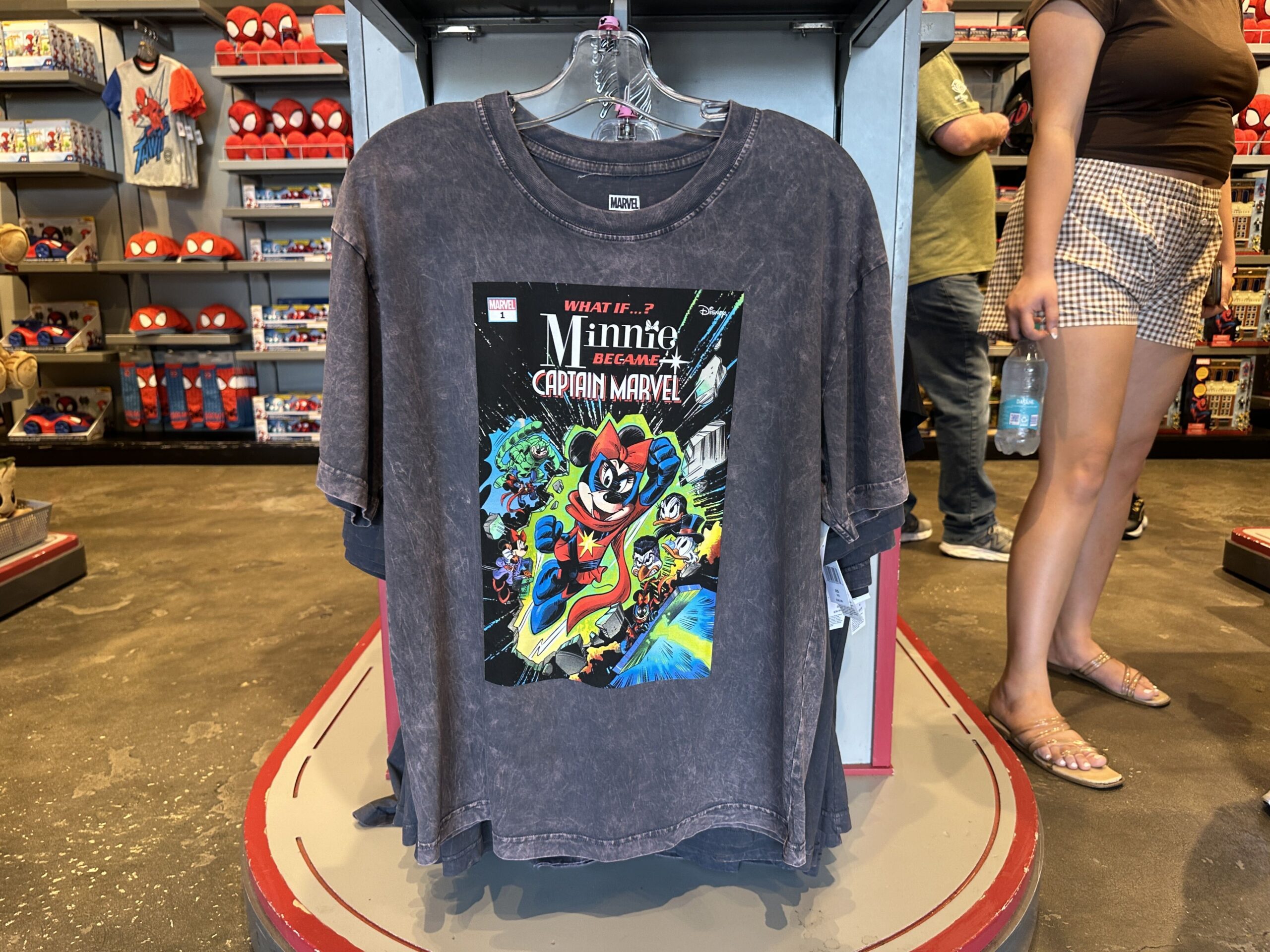 On a dark grey acid wash style t-shirt is a "What if...?" comic book cover of Minnie Mouse as Captain Marvel. She is seen flying through space with other characters behind her, like Scrooge McDuck and Magica De Spell.