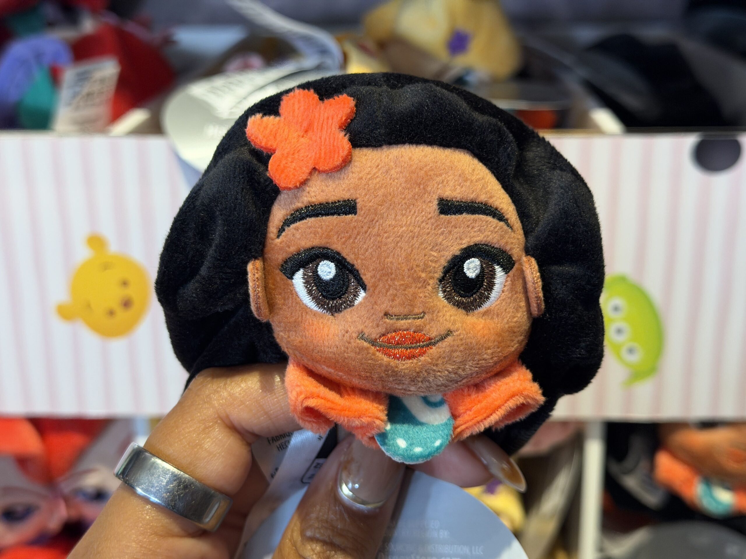 Moana's plush also has a fabric ruffle beneath her face, but her orange ruffle has her necklace pendant (the Heart of Te Fiti) in the center. There is an orange flower on the right side of her hair. All of Moana's facial features are embroidered.