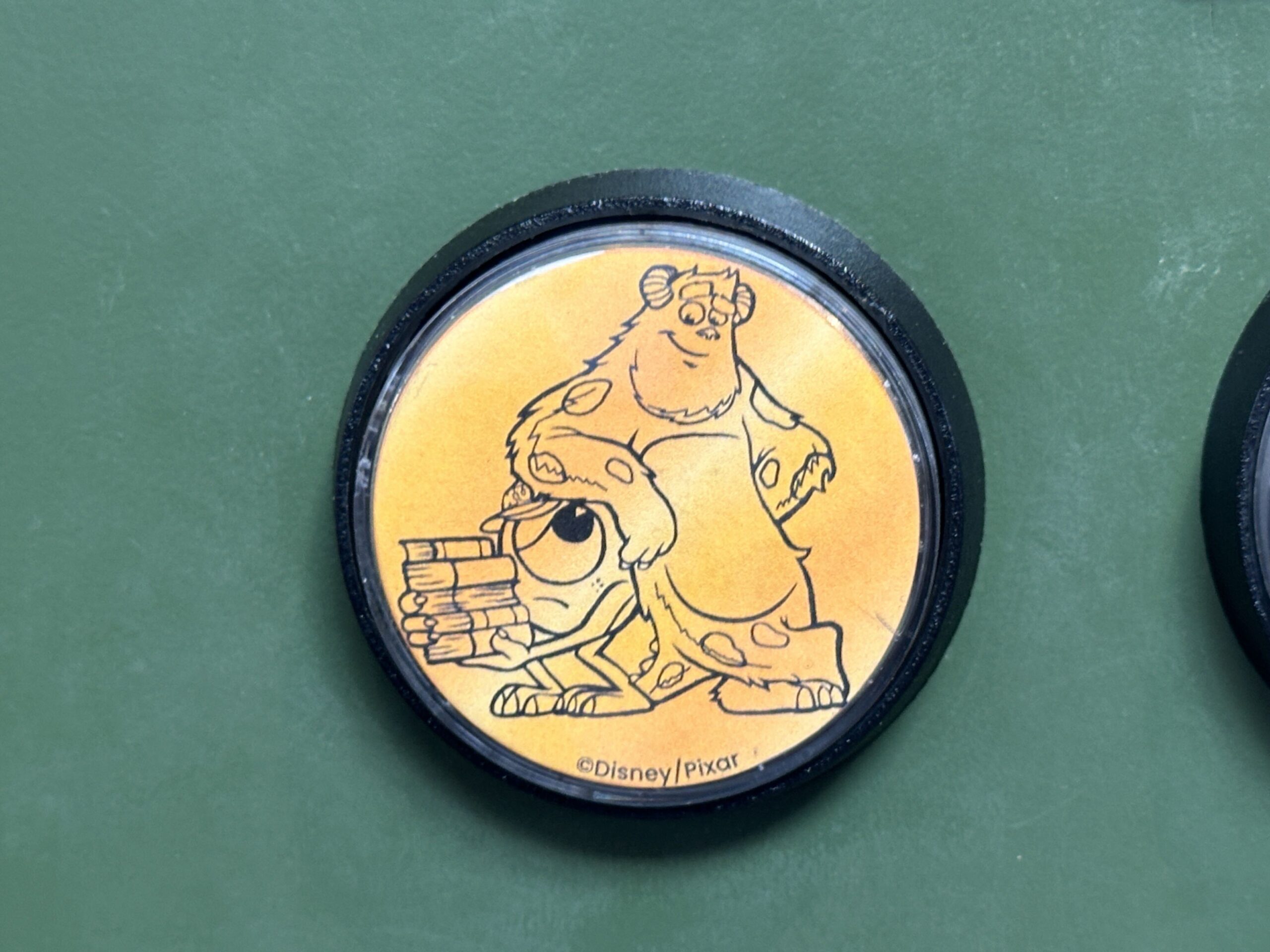 These collectible gold-tone medallions show Sulley leaning on Mike. Mike is wearing his Monsters University baseball cap and carrying a stack of books.
