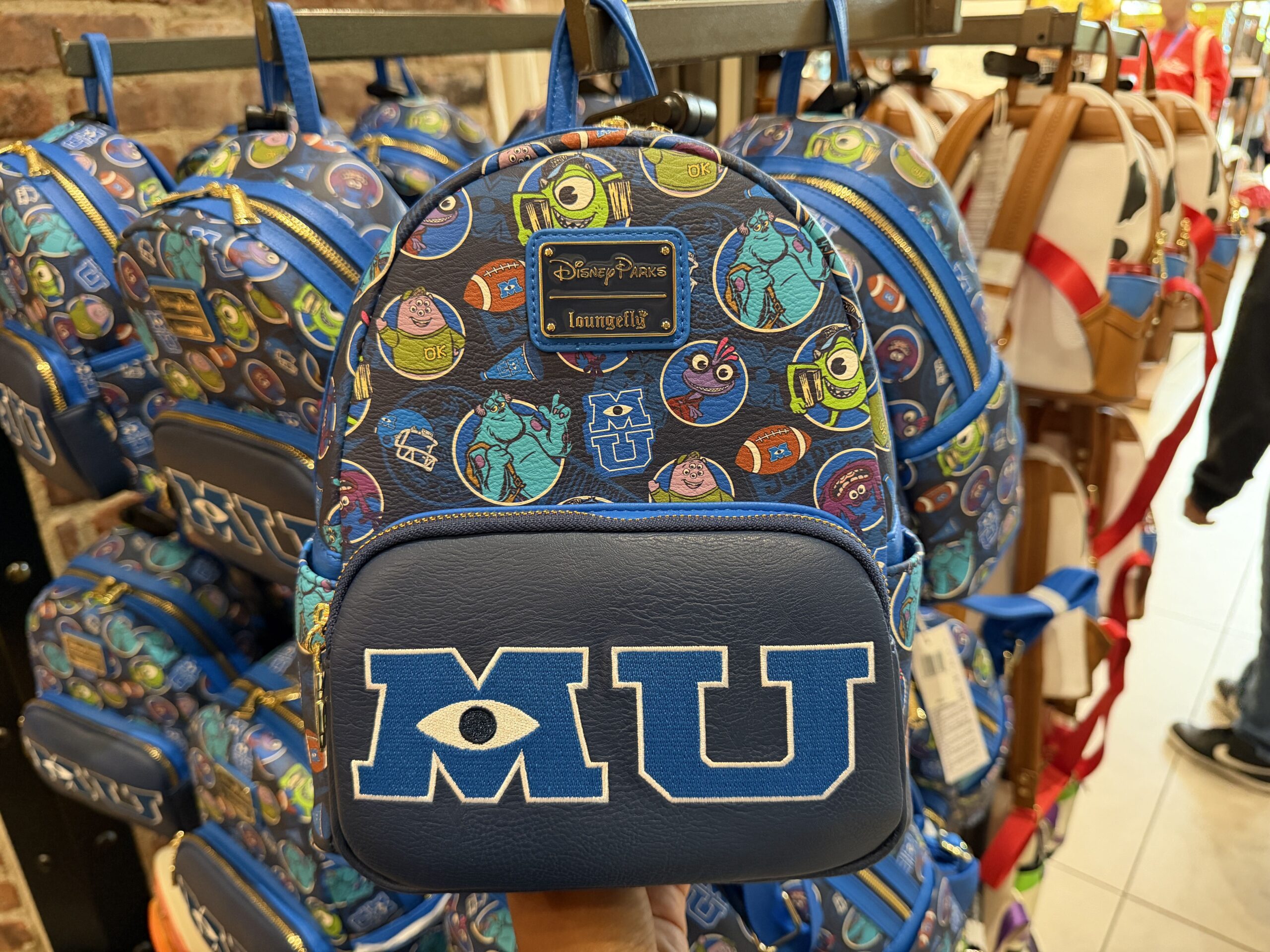 In blue, this mini backpack is covered in characters and iconography from Monsters University. We see returning characters like Sulley, Mike Wazowski, and Randall as university students, as well as new characters Squishy and Art. The front zippered pocket has the school's "MU" logo with a matching zipper pull.