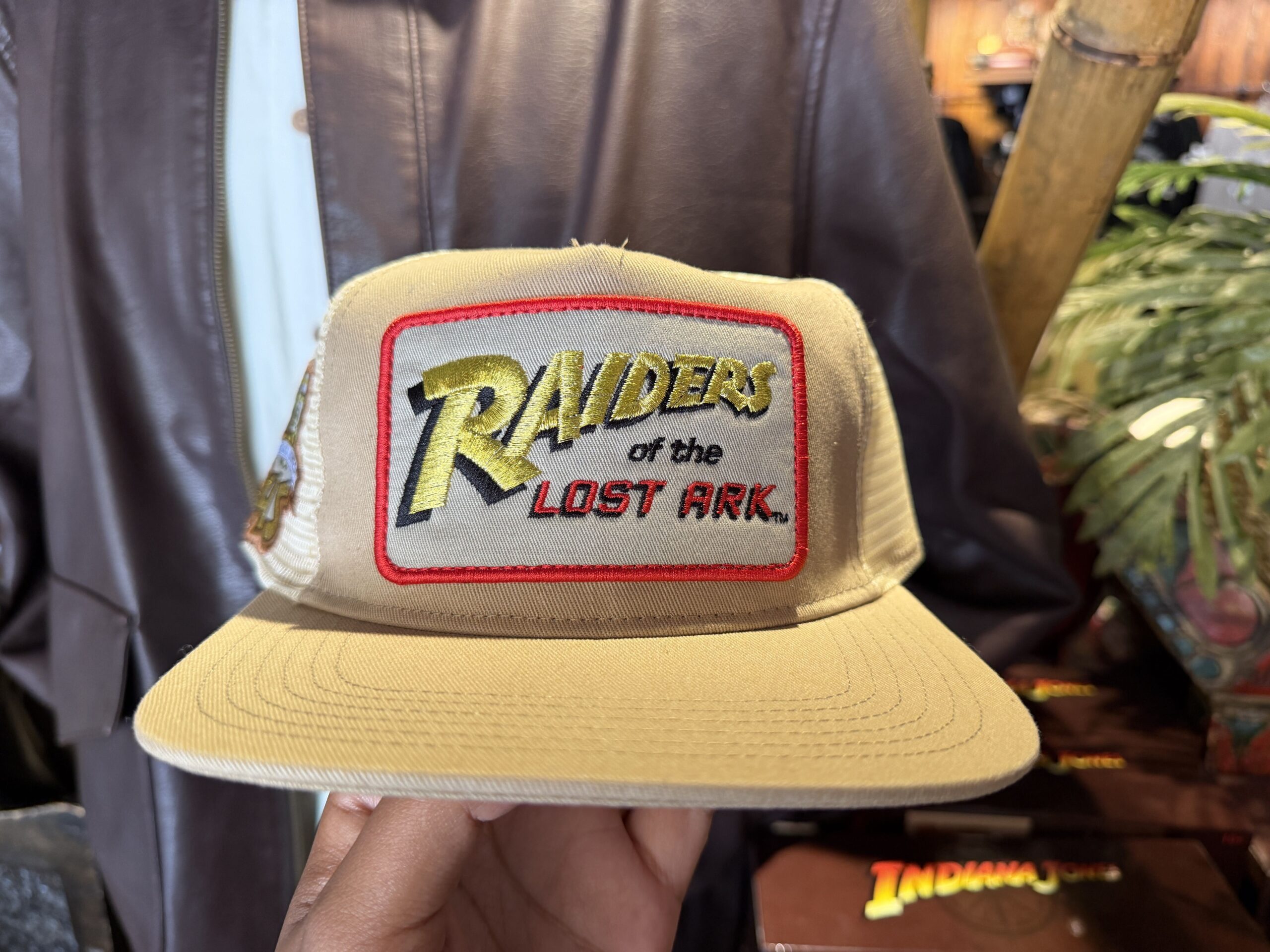 This tan flat-brimmed cap has a patch of the Raiders of the Lost Ark logo on the front. While the front of the cap is a solid fabric, the rest of the cap is made with a similarly colored mesh material.