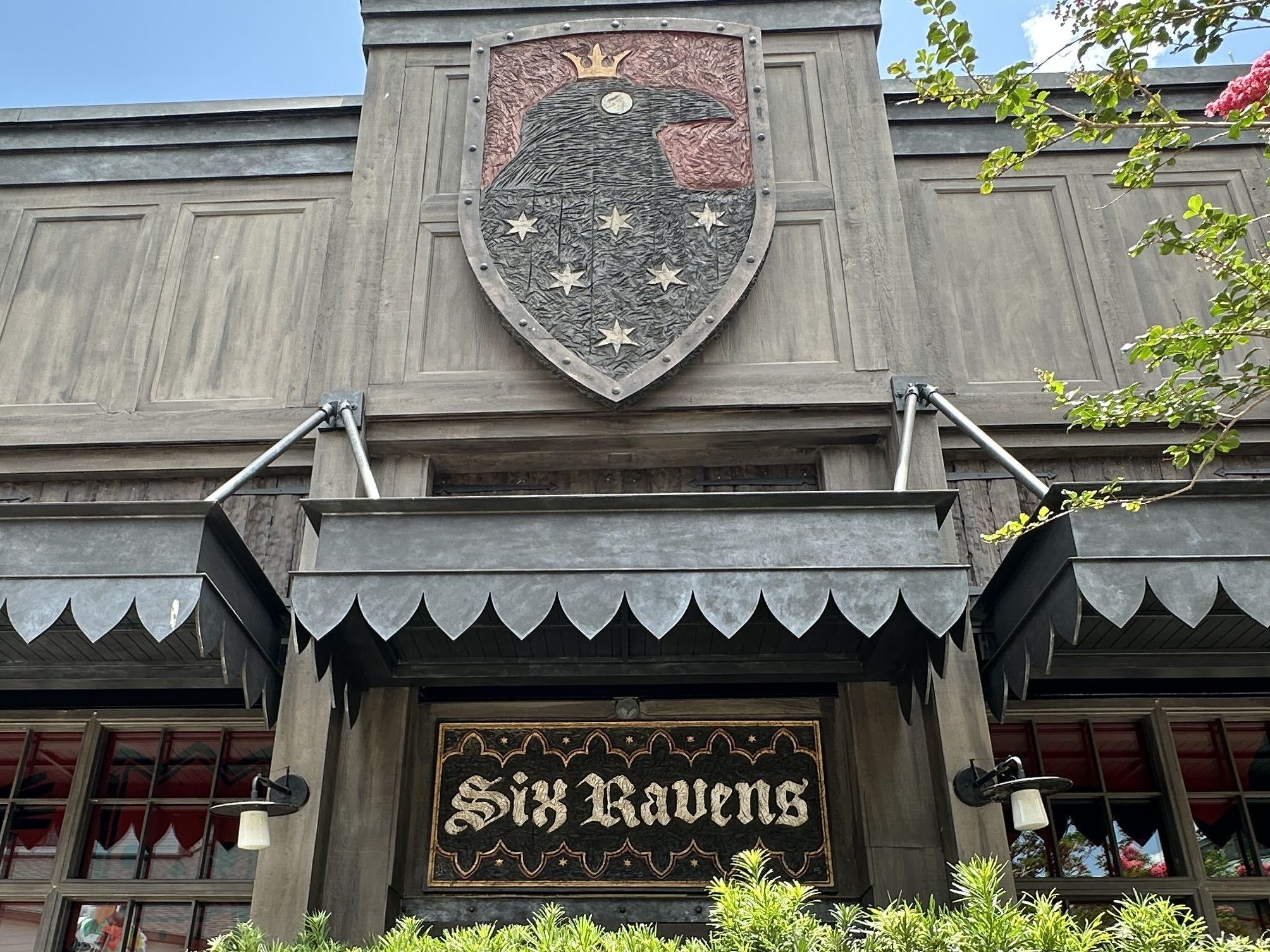 The logo sign above the awning is a shield shape with a raven. The raven is wearing a gold crown with six white stars on its body. Beneath the awning is a similarly themed sign that reads "Six Ravens" in a Medieval-style font.