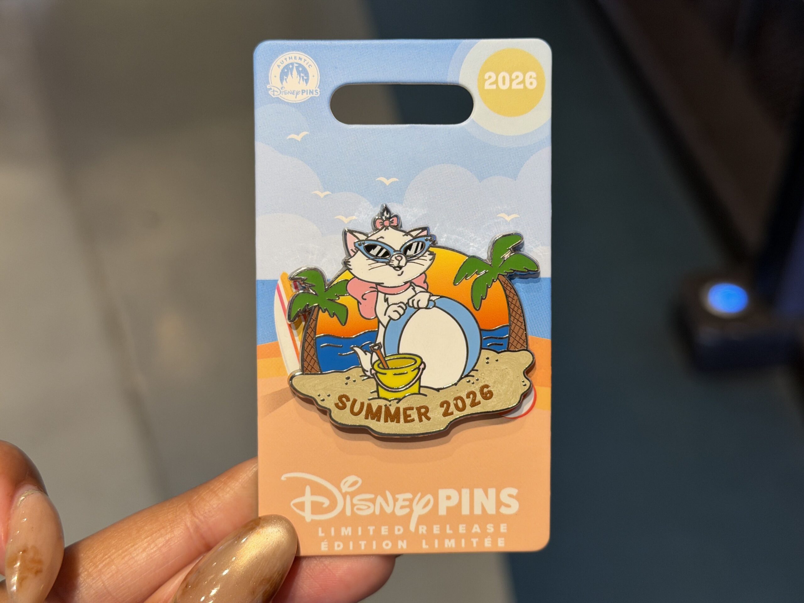 This pin shows Marie in her signature bow, wearing blue cat-eye sunglasses. She is standing with an oversized beachball on the beach. The ocean is behind her, and the pin is flanked by a palm tree on each side. Beneath the beach ball and sand pail is “Summer 2026” written in the sand.