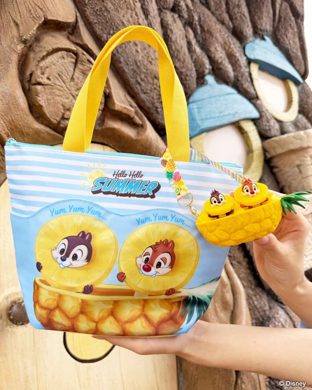 There is also a Souvenir Lunch Case in the same style. It has a Chip n Dale section pocket for small items. The souvenir case has blue and white stripes with an image of Chip n Dale eating a slice of pineapple as they sit in a boat of the fruit. Text at the top of the bag reads, "Hello Hello Summer," and the top handles are yellow.