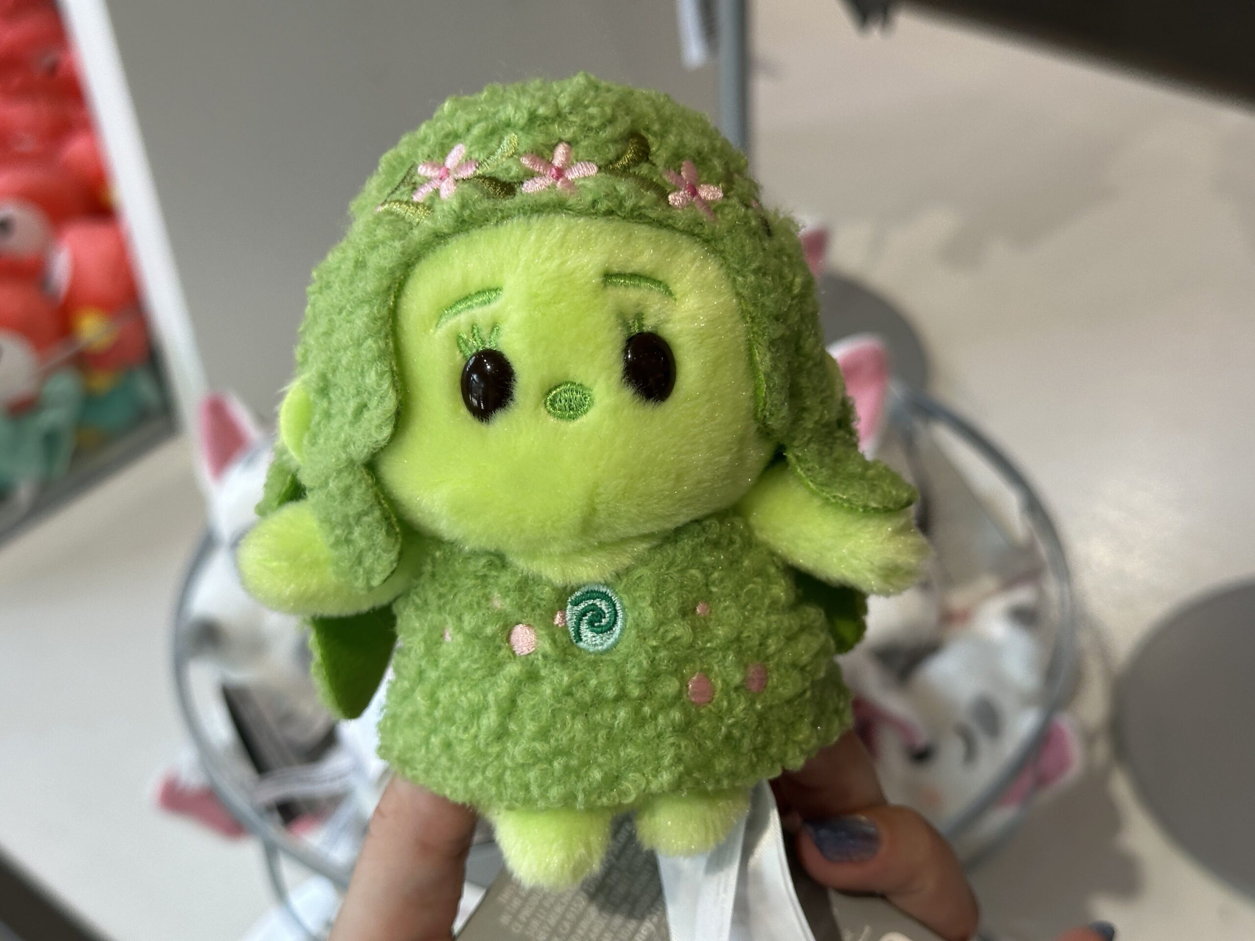 A more rarely seen character in Moana merchandise, Te Fiti, is shown in a standing position. Her hair and dress are a matching tufted material. Her heart, nose, eyelashes, and eyebrows are embroidered. So too are the pink flowers with green leaves or petals shown throughout her hair and dress.