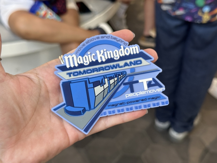The Tomorrowland PeopleMover magnet features the park's Magic Kingdom text logo in white with Tomorrowland written below in blue. "TTA peoplemover magnet-powered magic" is to the right of a ride vehicle. "Move above and beyond" is written along the top of the magnet.