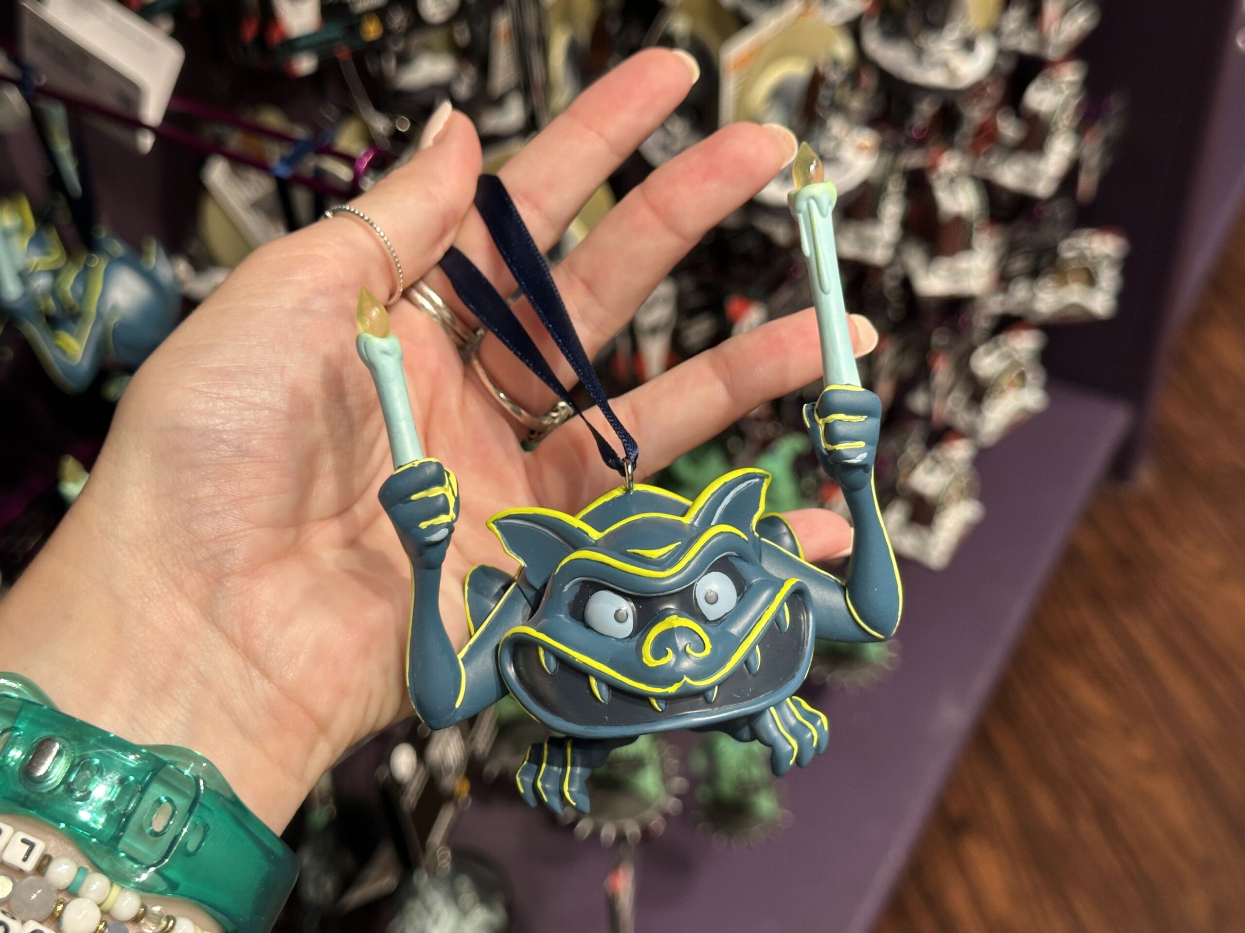 The gargoyle ornament similarly has yellow highlights against its blue-green exterior. It has a wide toothy mouth and sits in a crouched position. Two hands hold up a pair of candles.