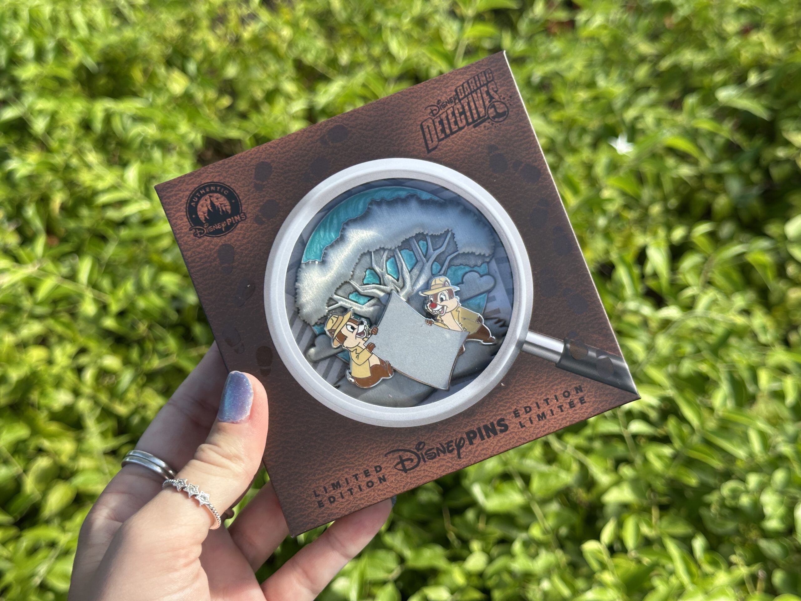 The Animal Kingdom mini jumbo pin features Chip 'n' Dale in detective outfits with a big piece of paper between them. The Tree of Life is in silver relief behind them, against a blue background. The packaging resembles a magnifying glass on brown leather.