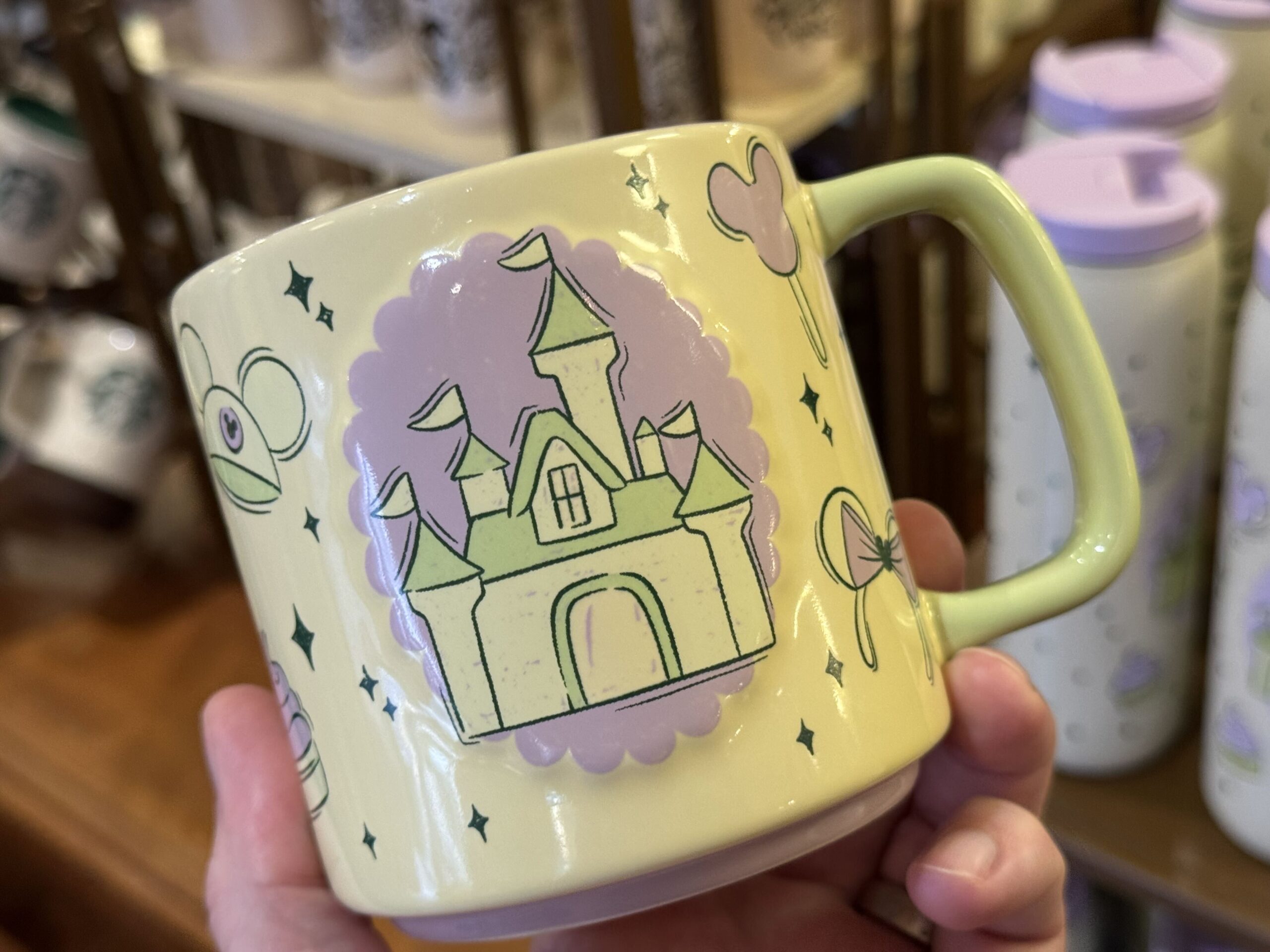 The mug is pale yellow with a lime green handle. Yellow, green, and lilac designs across the sides include a Fantasyland castle, Mickey ear hat, ear headband, and iconic treats. Dark green four-pointed stars accent the images.
