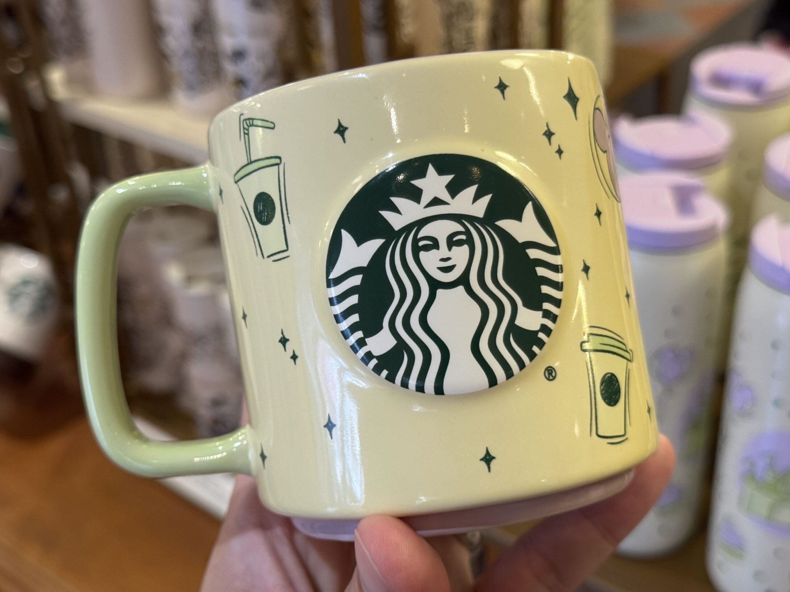 The mug is pale yellow with a lime green handle. Yellow, green, and lilac designs across the sides include a Fantasyland castle, Mickey ear hat, ear headband, and iconic treats. Dark green four-pointed stars accent the images.
