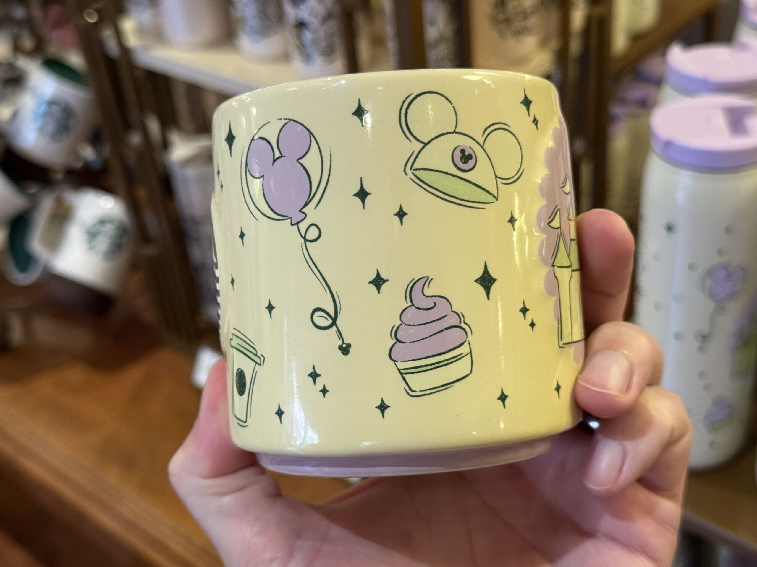 The mug is pale yellow with a lime green handle. Yellow, green, and lilac designs across the sides include a Fantasyland castle, Mickey ear hat, ear headband, and iconic treats. Dark green four-pointed stars accent the images.