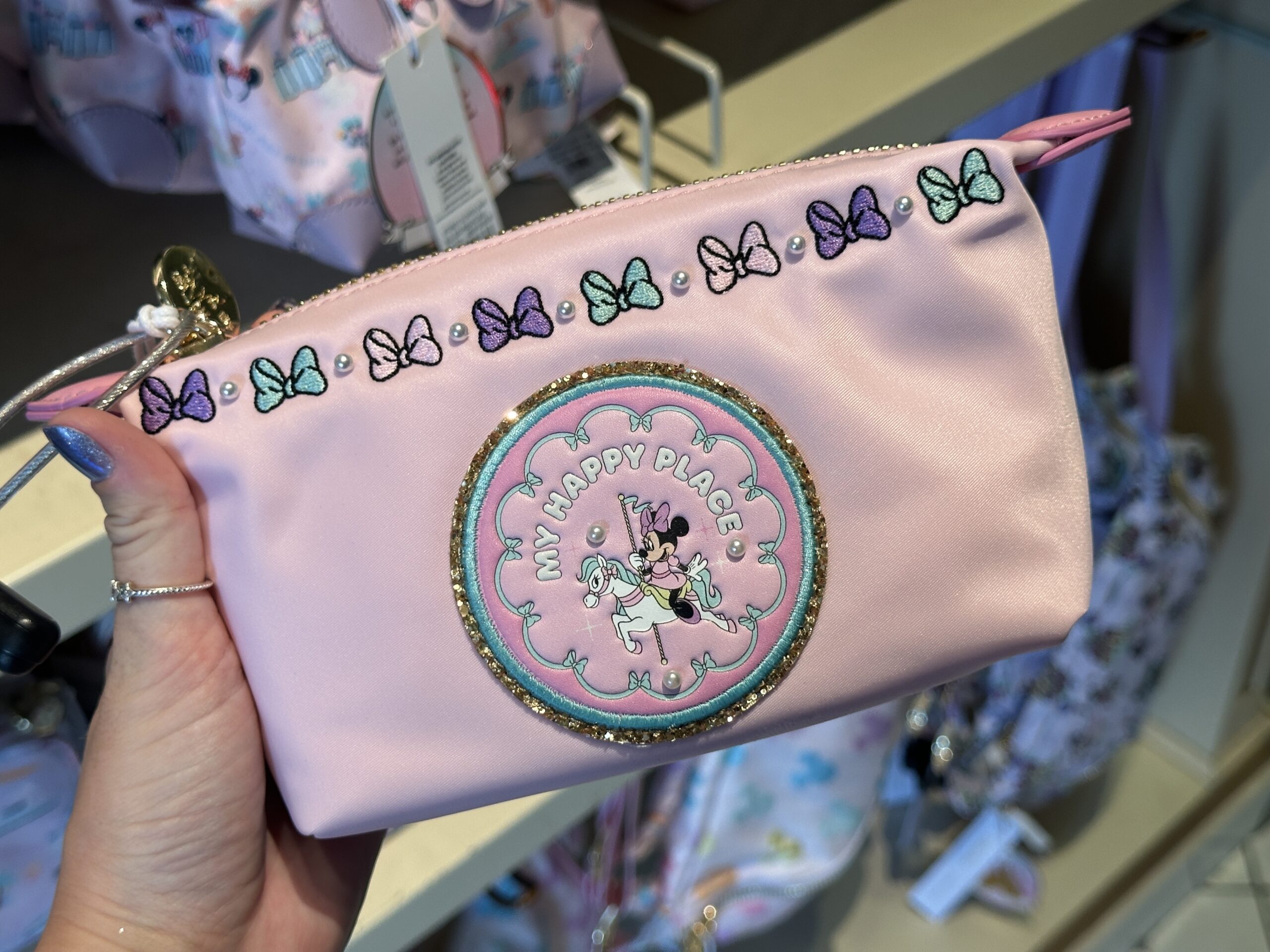 This faux leather zip pouch is adorned with a circular appliqué featuring Mickey on a carousel horse. Three pearls are around her with the words "My Happy Place." Above the patch are embroidered purple, aqua, and pink bows separated by pearls.
