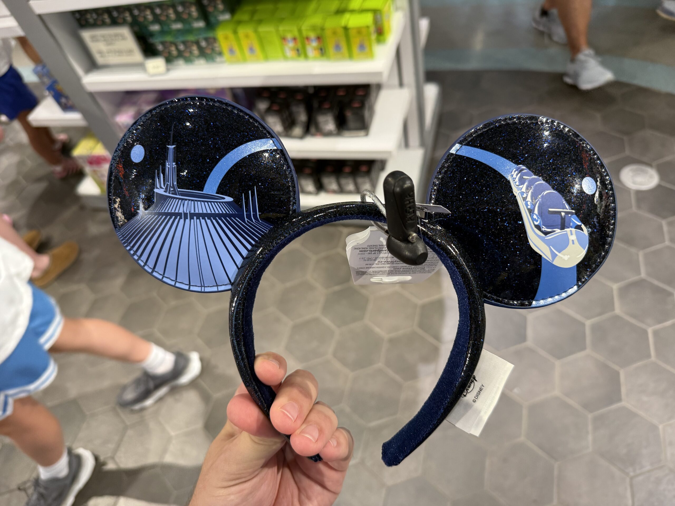 This ear headband is black and has a subtle sparkly sheen. The Space Mountain building and ride vehicle are depicted in blue on one side, while the ride vehicles from Astro Orbiter and Tomorrowland Speedway are on the other side.